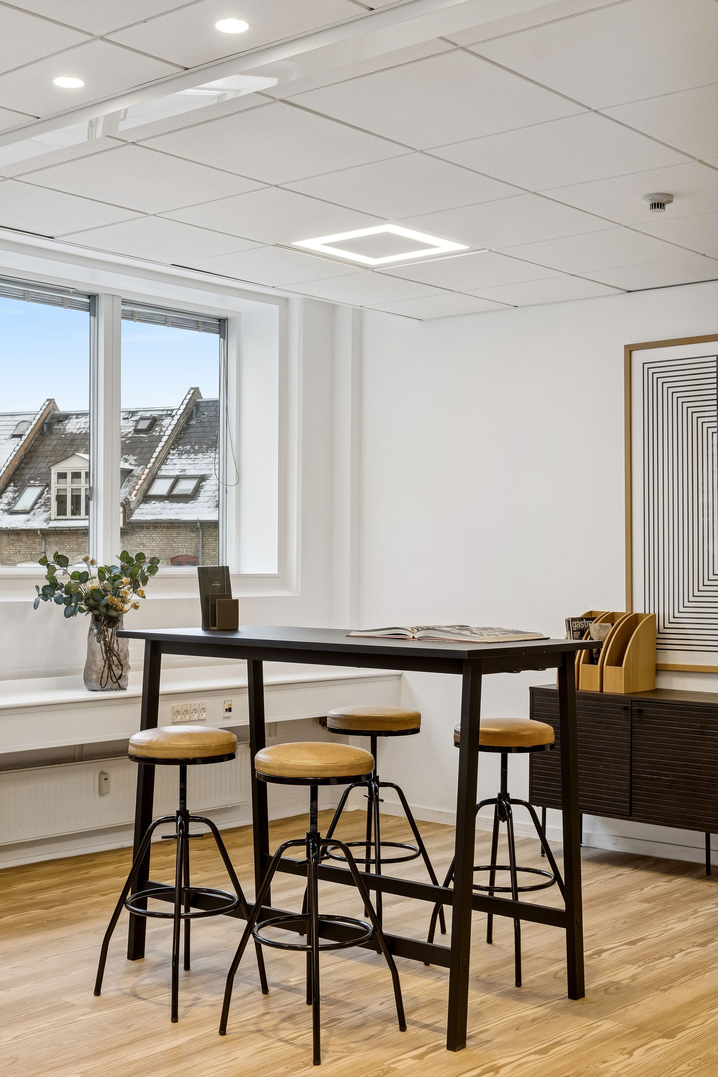 Kontor med lyse lofter, store ruder og trægulv. Rundt bord med fire lædersæder og plante i vase. Sort kabinet med papirholder og maleri i ramme på væggen.