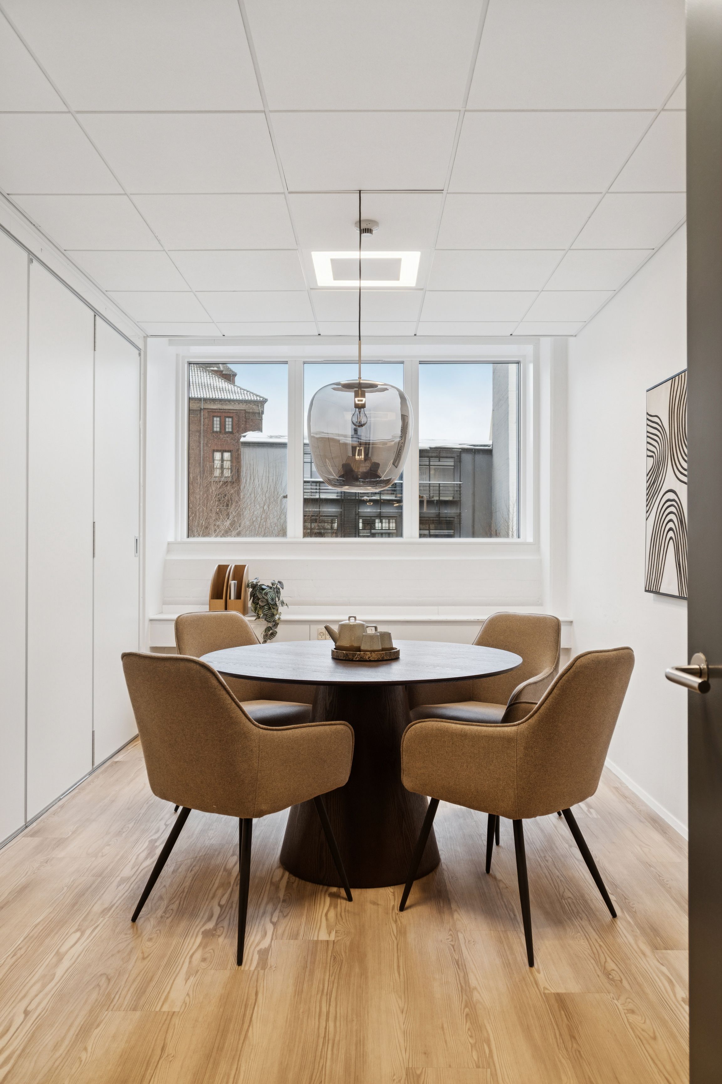 Moderne kontor med runde borde og stolpeformet loftslampe i lyst rum med trægulv og stort vindue