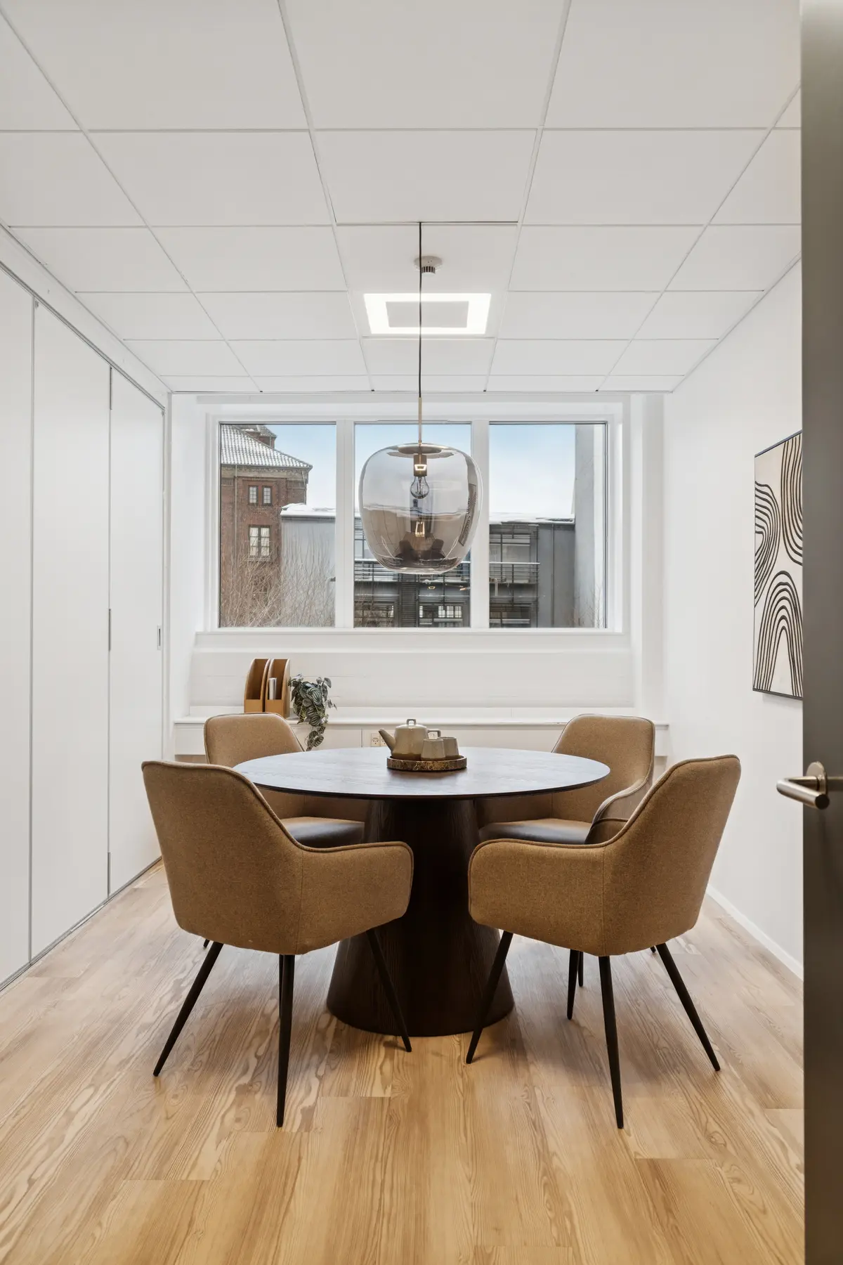 Moderne kontor med runde borde og stolpeformet loftslampe i lyst rum med trægulv og stort vindue