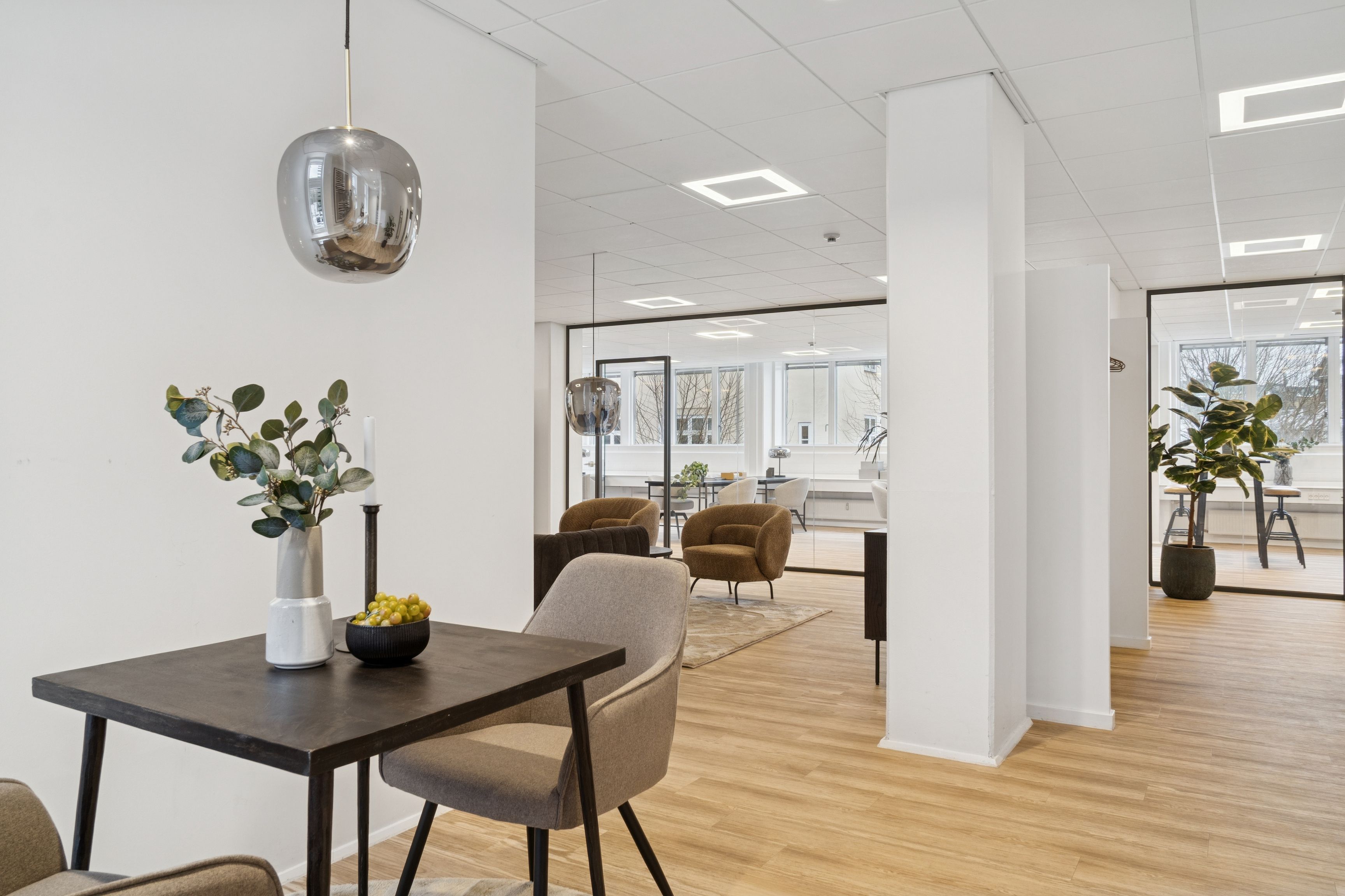 Kontor med lyse lofter, hvid væg og møbler i sort og beige, udsmykket med lysekroner og plante. Rundt om er der lyse gulve og glasvinduer med udsigt til byen