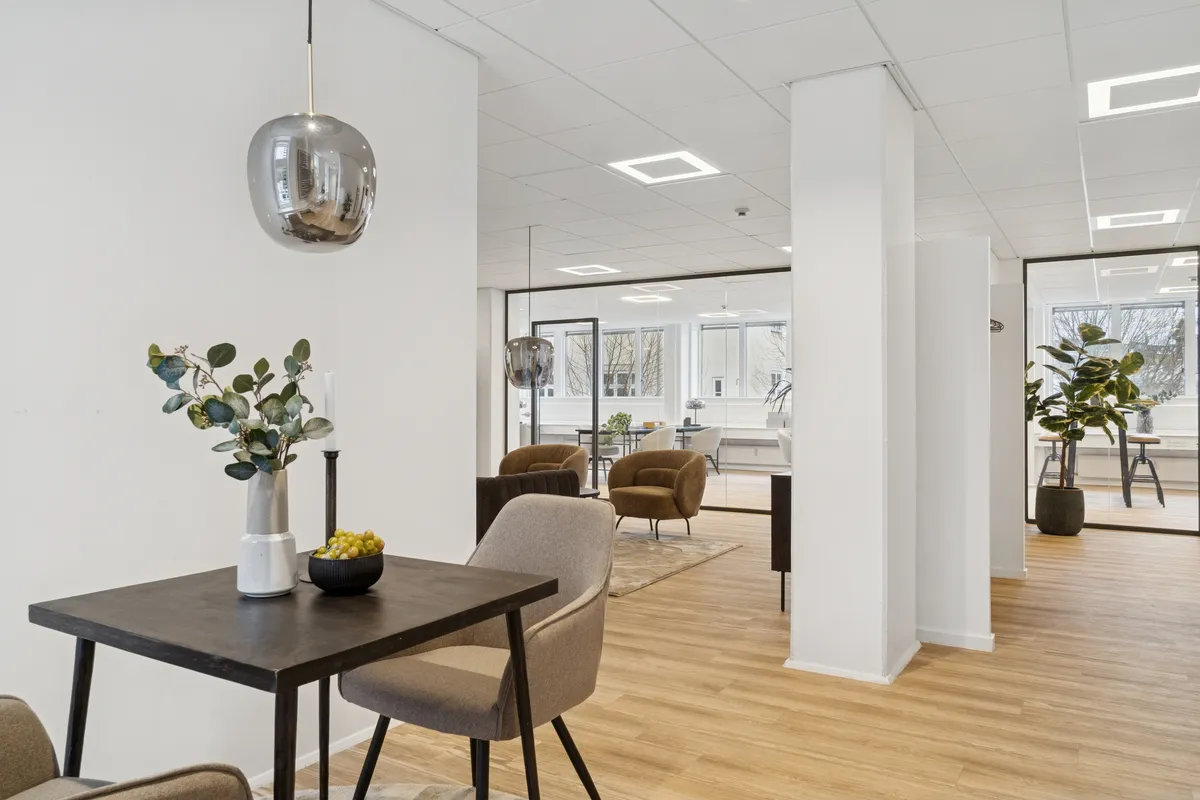 Kontor med lyse lofter, hvid væg og møbler i sort og beige, udsmykket med lysekroner og plante. Rundt om er der lyse gulve og glasvinduer med udsigt til byen