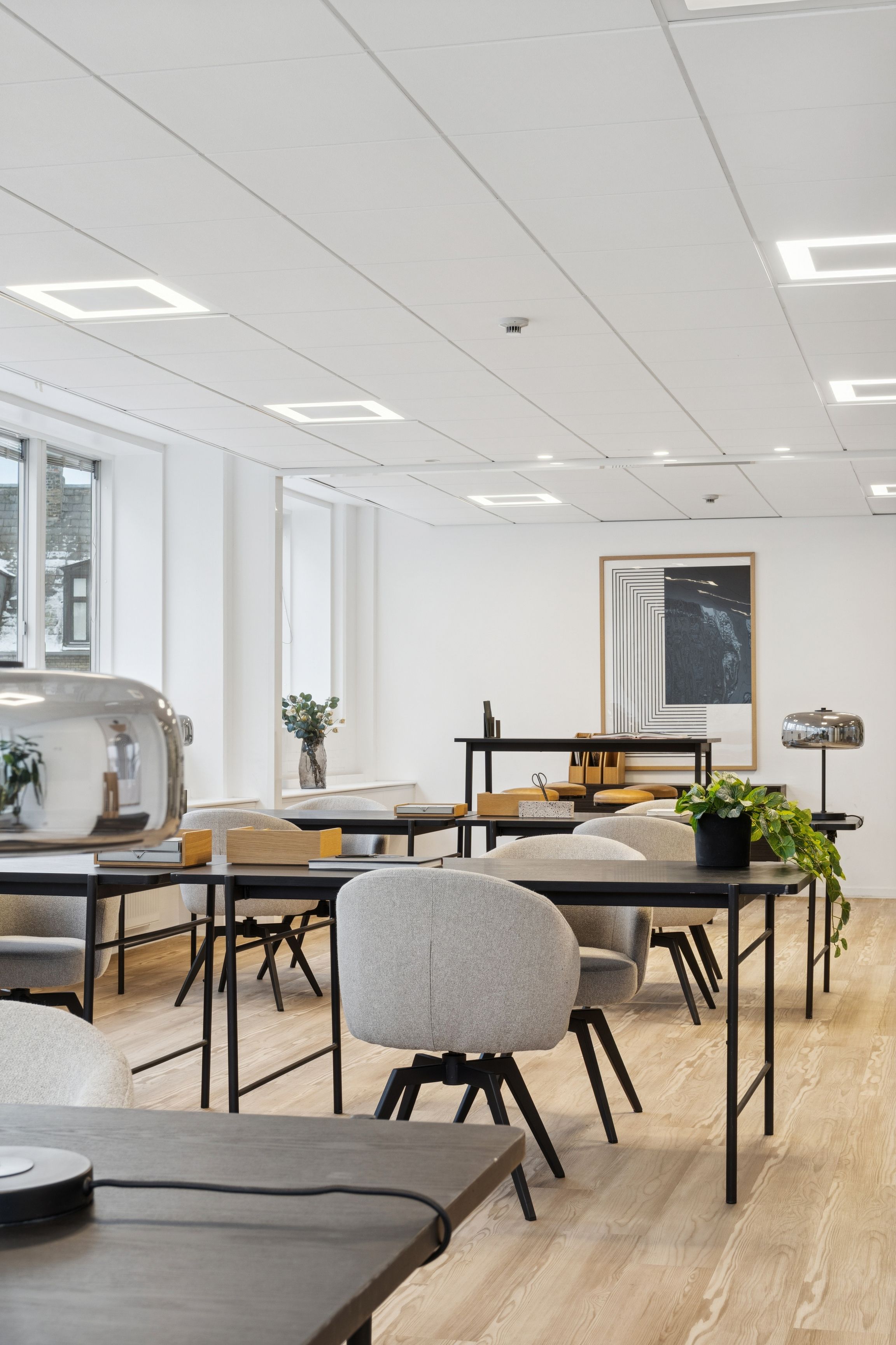 Moderne kontor med lyse gulve, hvide vægge og store vinduer mod Vesterbrogade. Kontorbord med skrivebord, stol og plante samt lyse loftslampe. Lyse firkantede loftsplader.