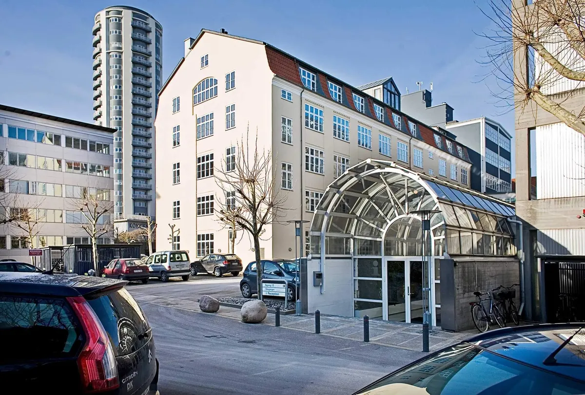 Bygning med glasdækket indgang og parkerede biler samt cykler i forgrunden og træer og høj bygning i baggrunden