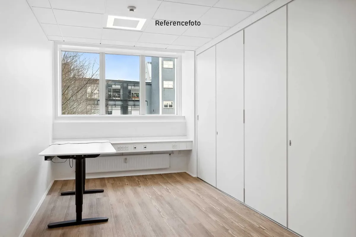Referencefoto af et tomt kontor med lyse hvide vægge og loft, højt vinduesglas og trægulv. Et skrivebord står mod en af