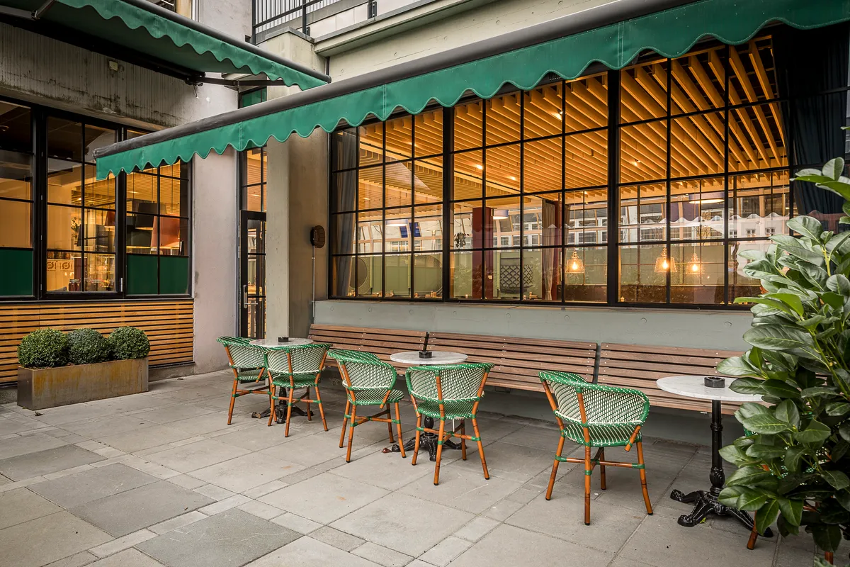 En terrasse med grønne stole og højrekantede borde foran et cafévindue med møbler og plantager