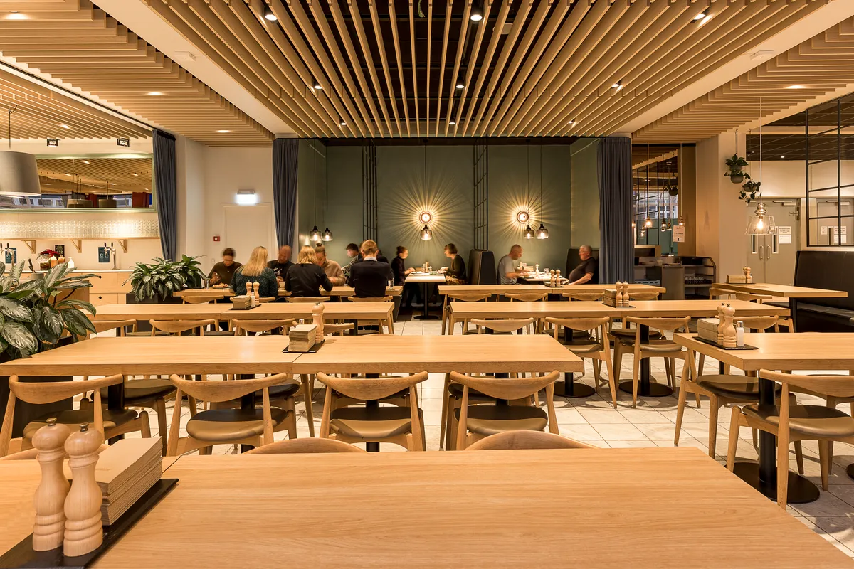 Billede af en moderne og rummelig restaurant med lyse træborde og stole, hvor gæsterne sidder og spiser, mens lyse lysekroner hænger fra loftet