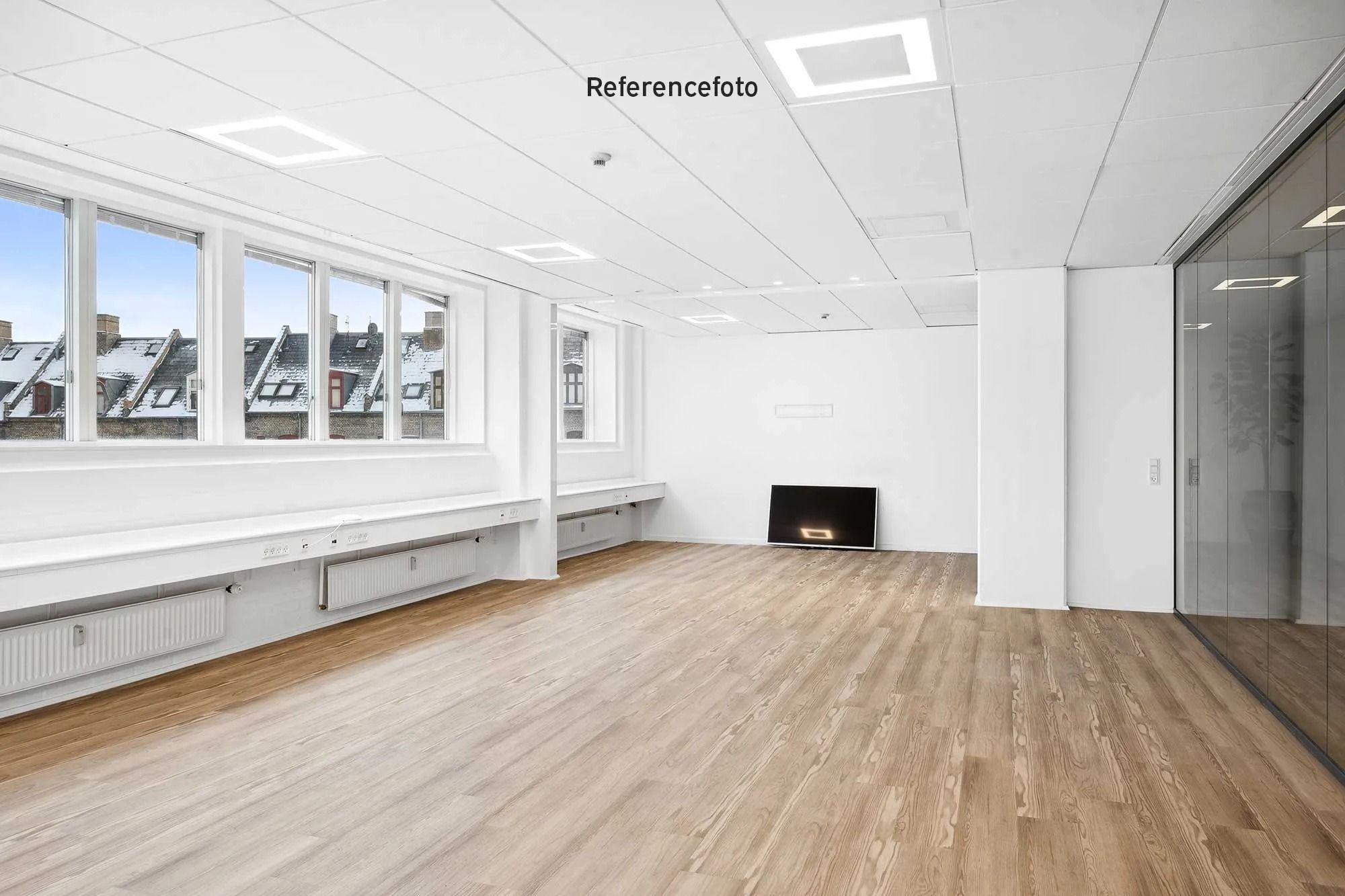 Referencefoto af en tom kontorrum med lysegrå vægge og loft, gulvtæppe og flere vinduer med udsigt til bygninger og huse med