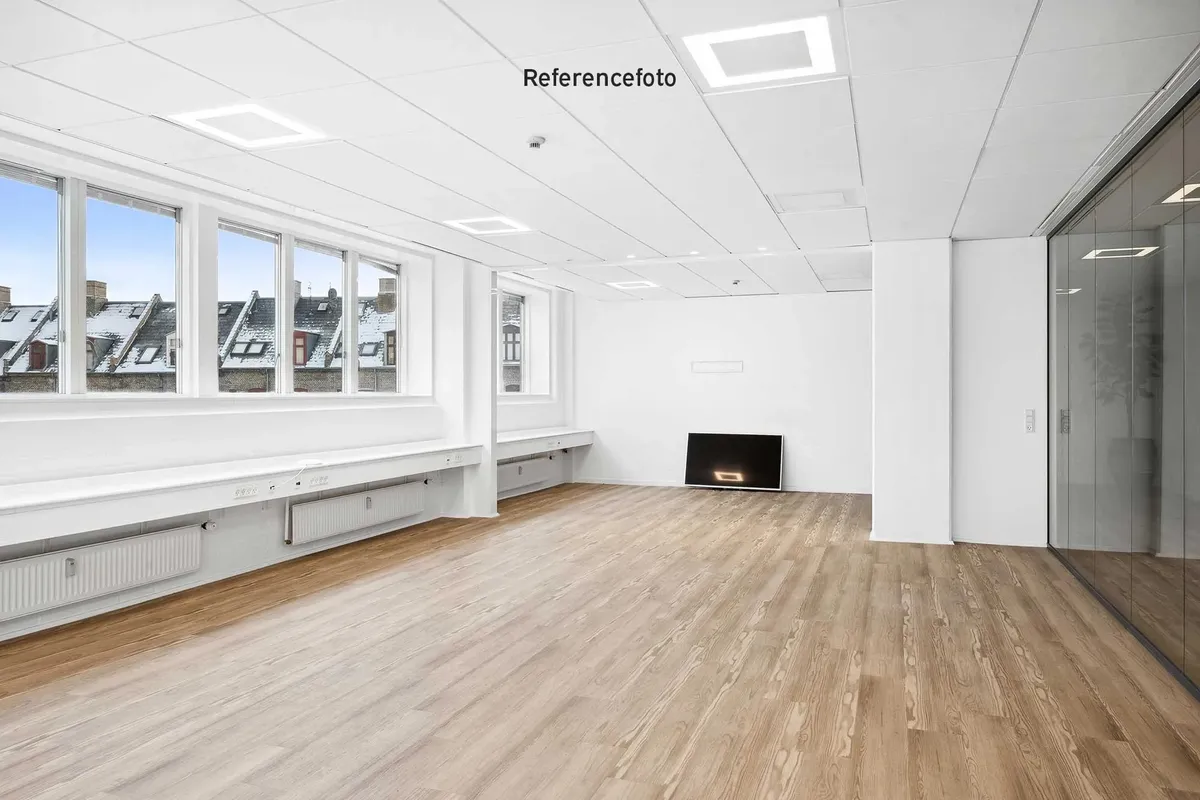 Referencefoto af en tom kontorrum med lysegrå vægge og loft, gulvtæppe og flere vinduer med udsigt til bygninger og huse med