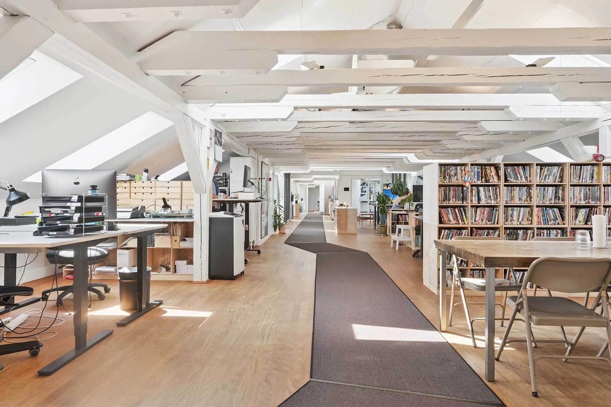 Moderne kontor med højloft og trægulv, to skriveborde med computere, to lænestol og et langt bord med en række hylder med