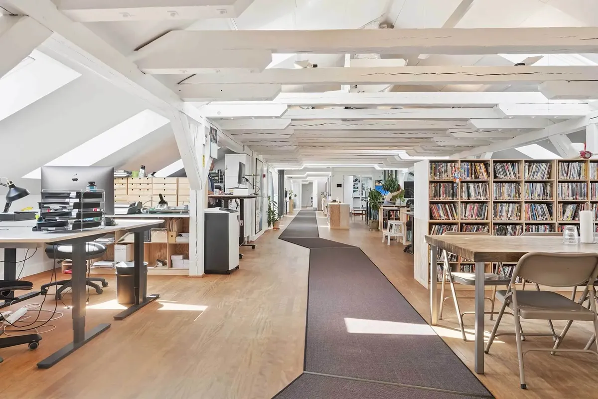 Moderne kontor med højloft og trægulv, to skriveborde med computere, to lænestol og et langt bord med en række hylder med