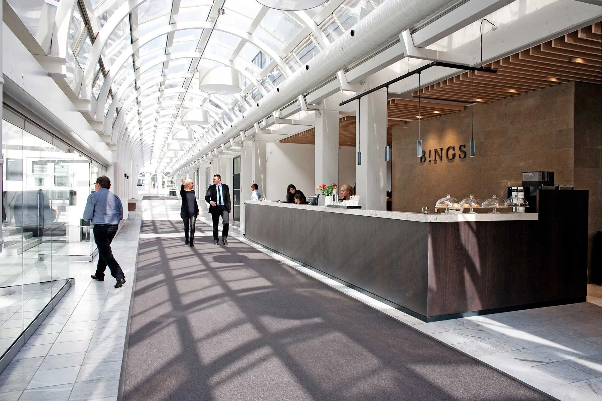 Modernt kontorhus med glasvinduer og søjler, flere mennesker går på gulvet, og en kvinde står ved baren