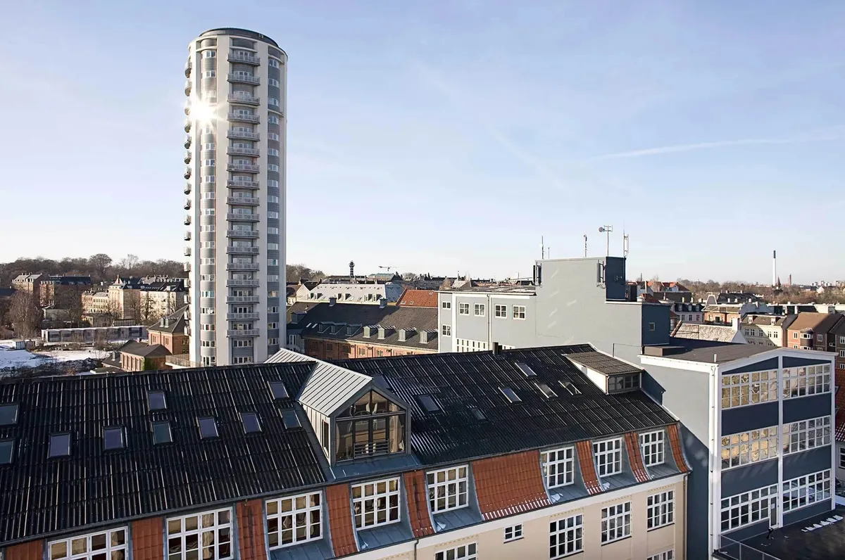 Moderne bygning med mange vinduer og en slank facade i baggrunden med flere bygninger og et solrigt, blaa himmel