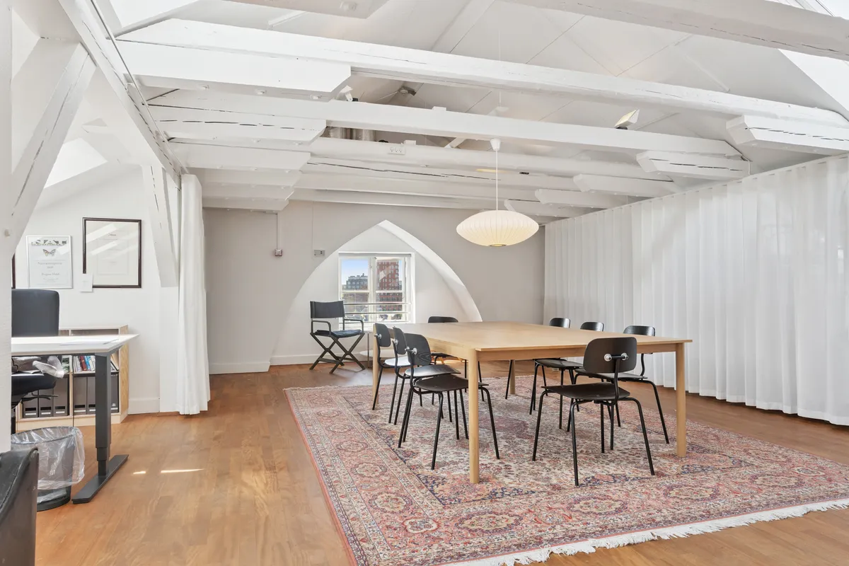 Kontor med lyse loftsbjælke, hvidt loft og gulvtæppe, moderne møbler og hvide gardiner, skrivebord og stol, vinduesbue med udsigt til byen