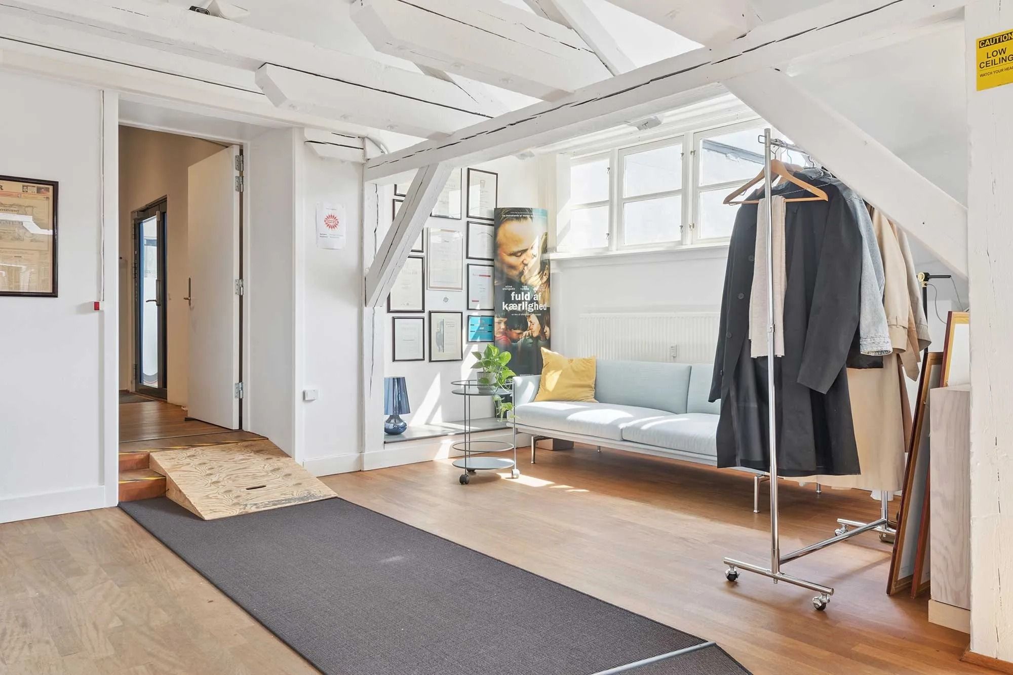 Lukket hvidt dørkarm med trappe i baggrunden og gulvbelægning i træ. En moderne, hvid sovesofa med pude i stuen. En