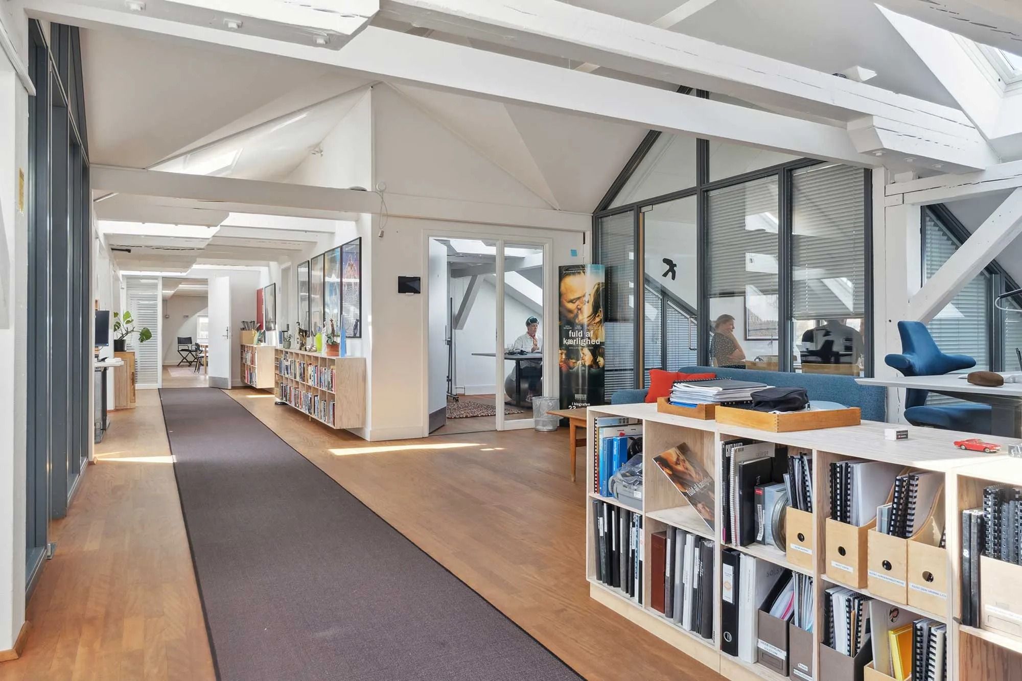 Moderne kontorrum med højt loft, lyse trægulve og hylder med bøger og papirer