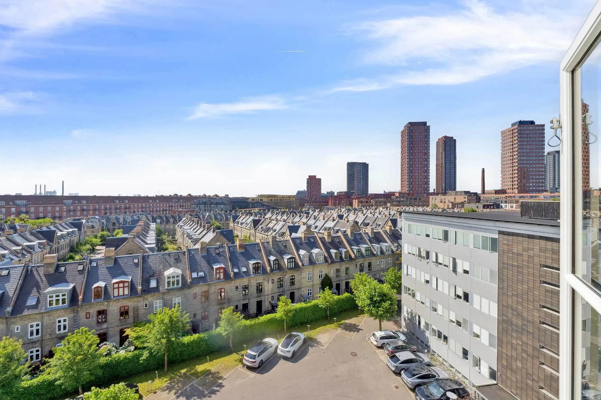 Moderne bygninger med parkeringsplads og grønne områder. En stor bygning med højderyg og flere høje bygninger i baggrunden.