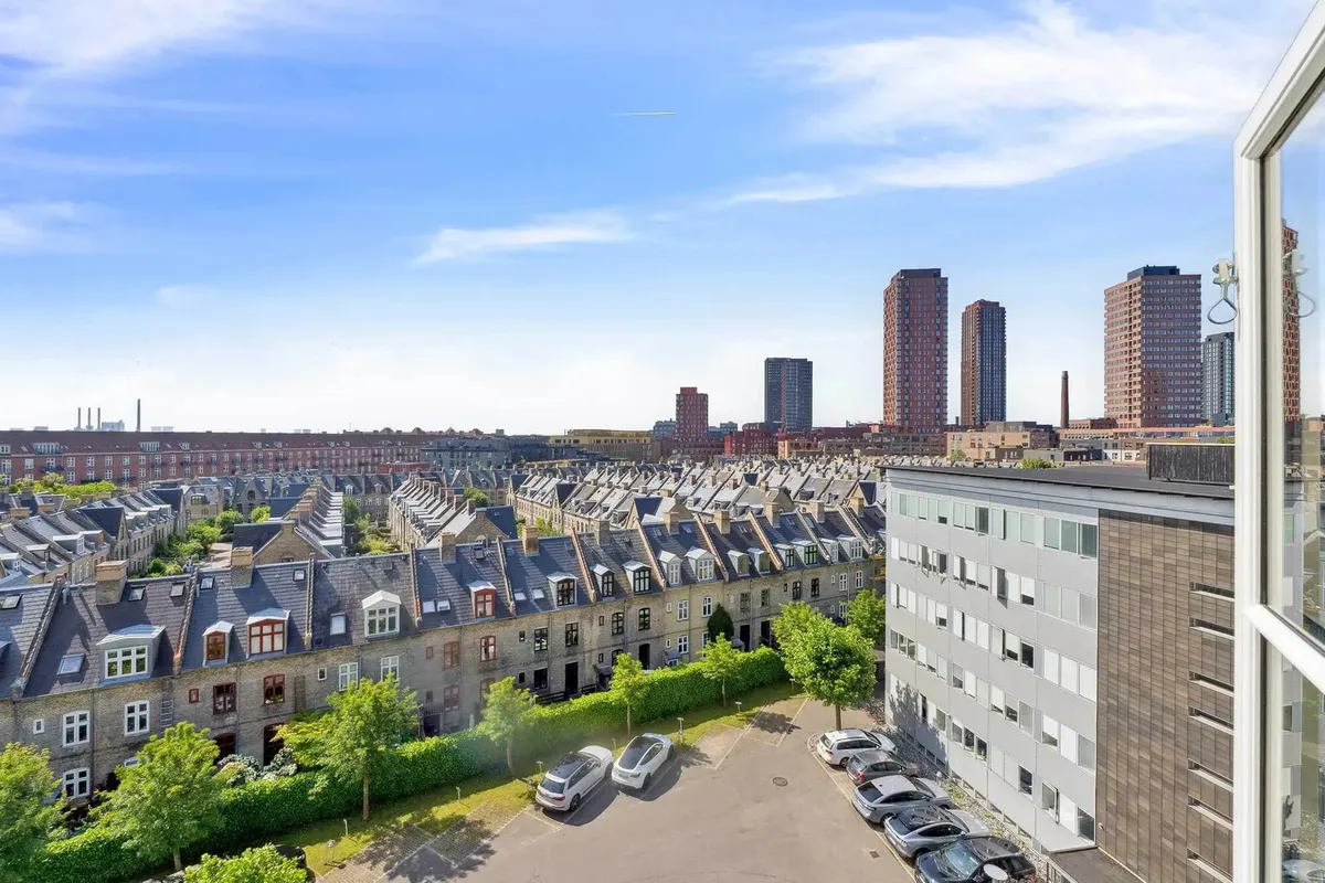 Moderne bygninger med parkeringsplads og grønne områder. En stor bygning med højderyg og flere høje bygninger i baggrunden.