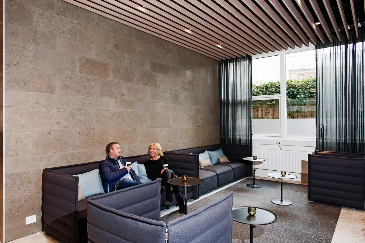 To mennesker sidder på lædersofaer og drikker kaffe ved et vindue i et kontor med lysegule vægge og møbler