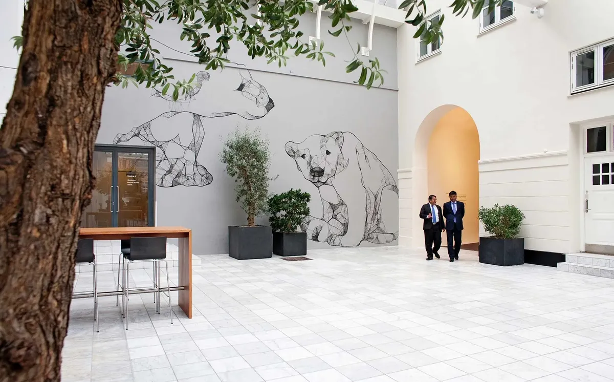 To forretningsmænd vandrer gennem gårdhave med gulv af marmor og kunstværk på væggen med to hunde og træer