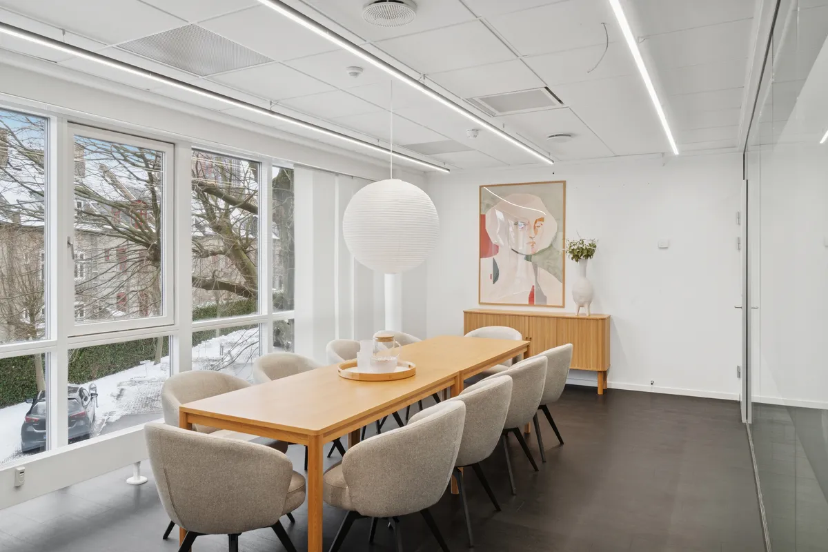 En lyse og moderne kontorrum med lyse vinduer og store ruder, hvor der er en lang træbord med stole og en hvid kugleskærm