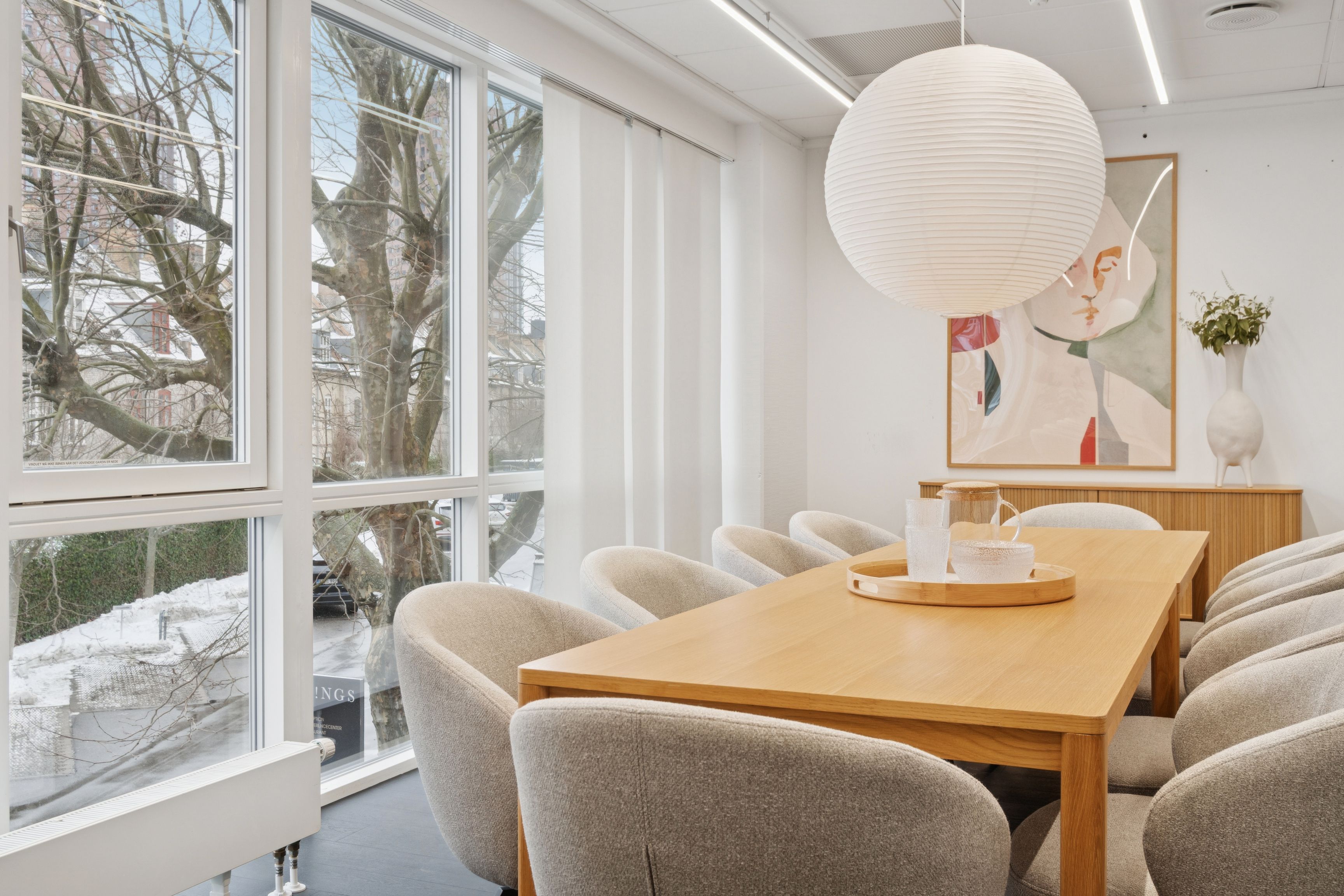 Kontor med vinduer med udsigt til sne, lyse loftslampe, træbord, beige stole, plante i vase og maleri på væggen