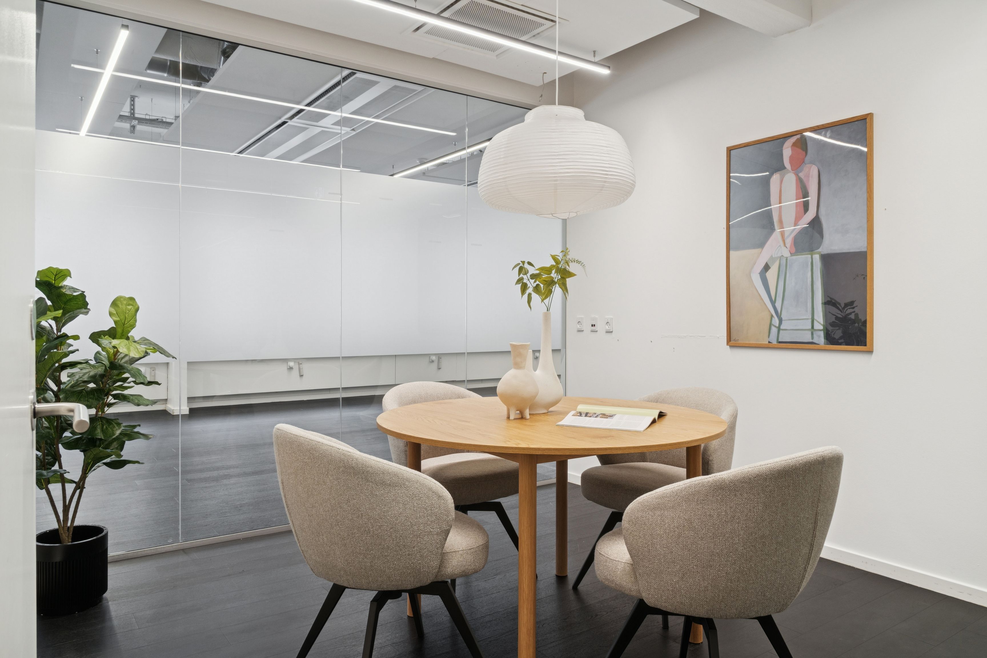 Minimalistisk kontor med rundt bord, høj loftshøjde og moderne møbler. En vase med en plante og et maleri på væggen. Lampe med hvidt kuppelloft.