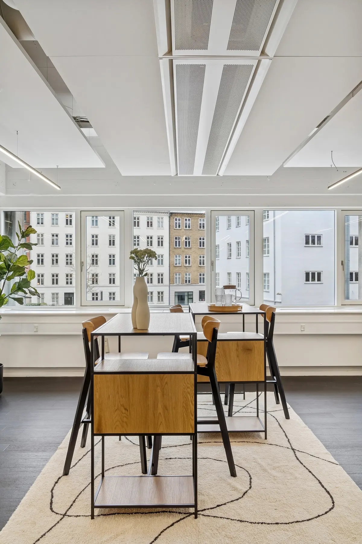 Moderne kontor med lyse vinduer og beige gulv. To borde med træ sæder og høje ben står på en beige tæppe med snoede linjer. Et hvidt kunstværk står på et bord ved vinduet