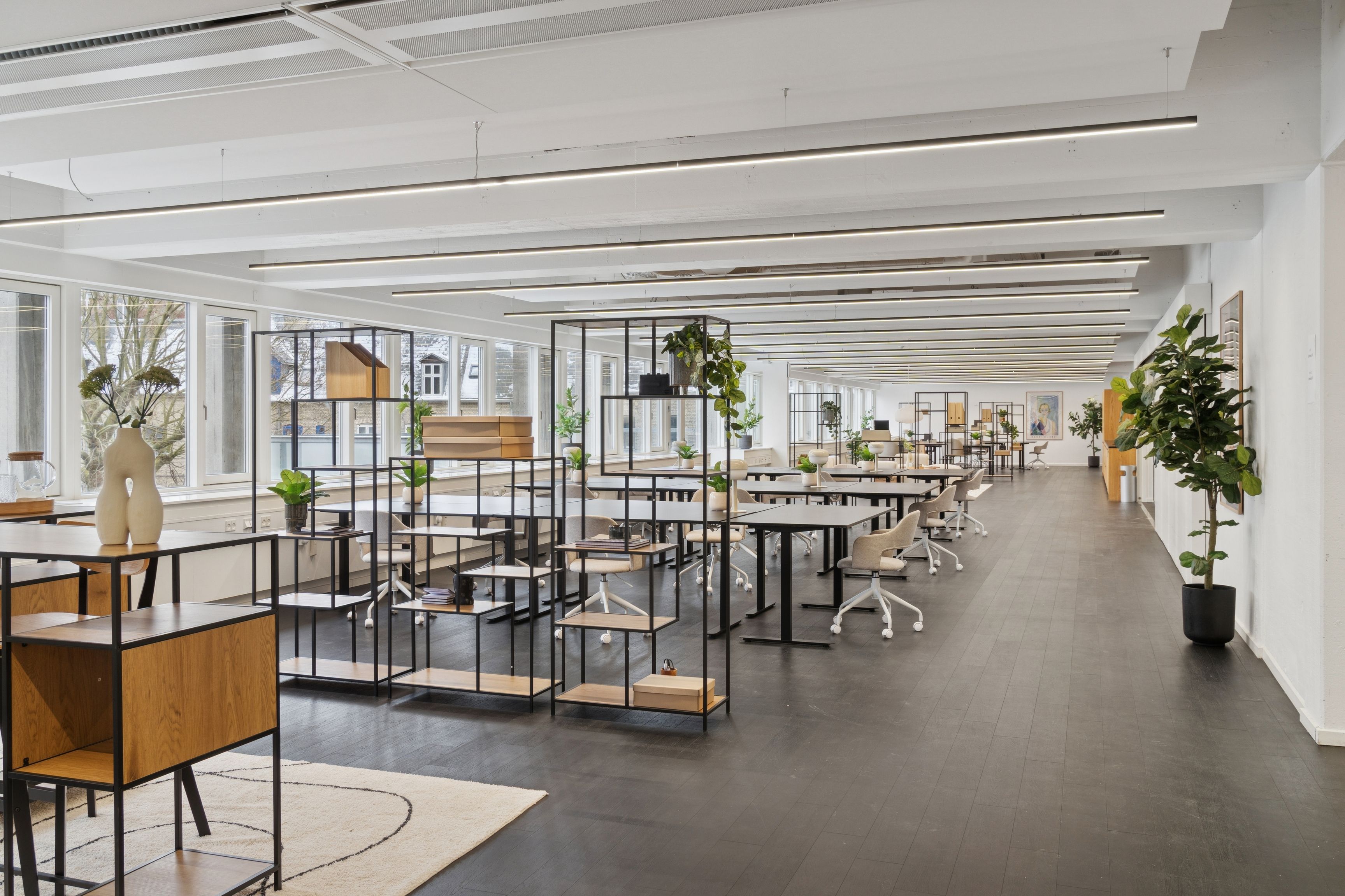 En stor lys kontorlokal med lysegult lys, hvide vægge og lysegrå gulv. Loftet er hvidt med lange, slanke, hvide lysrør. Væggen er dekoreret med flere små kunstværker og plantepotter