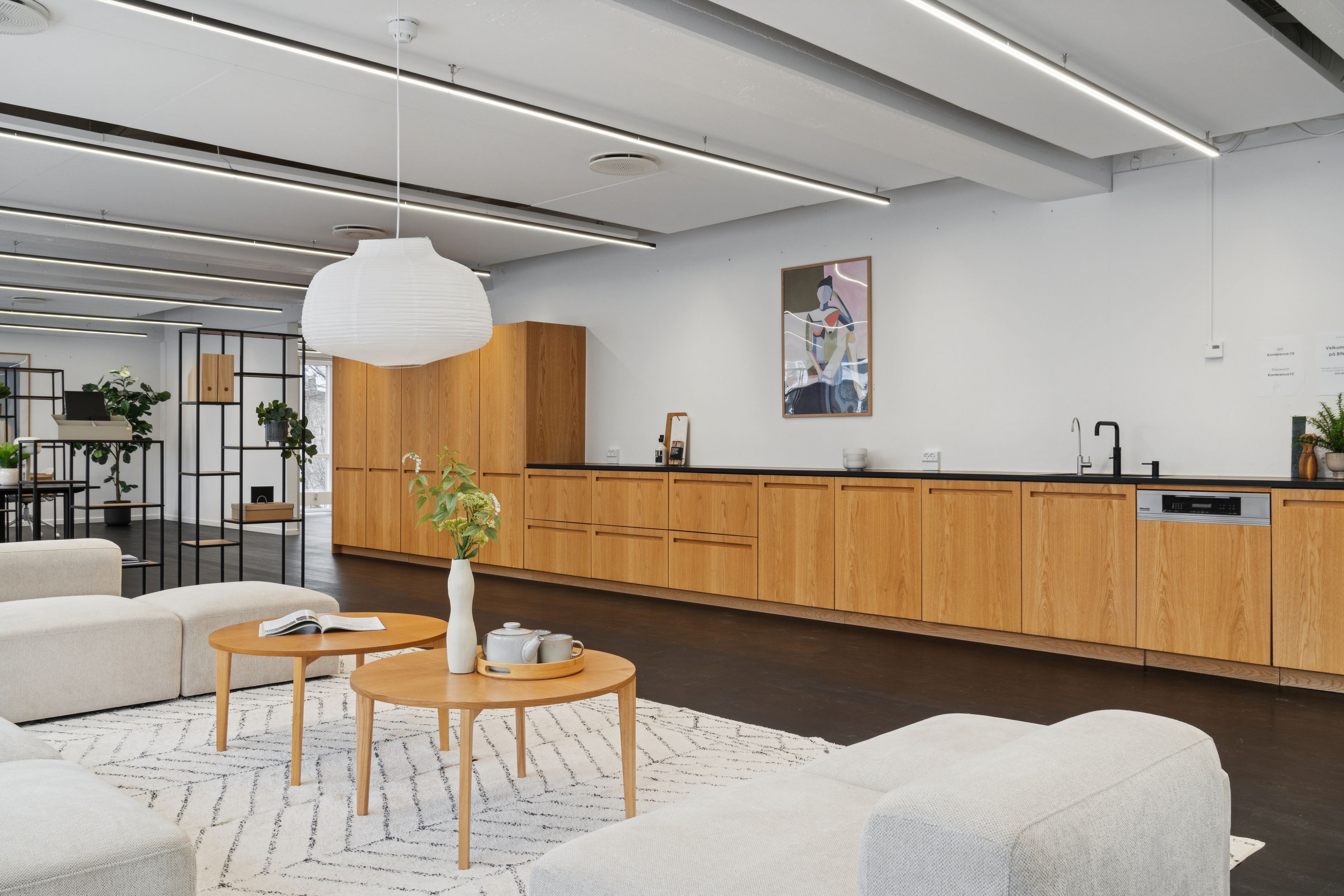 Kontor med åbent, moderne design og lyse farver. To bord med plante og tavle. Køkken med moderne møbler og lyse loftslampe. Sofas og hjørnebord i lys grå farve