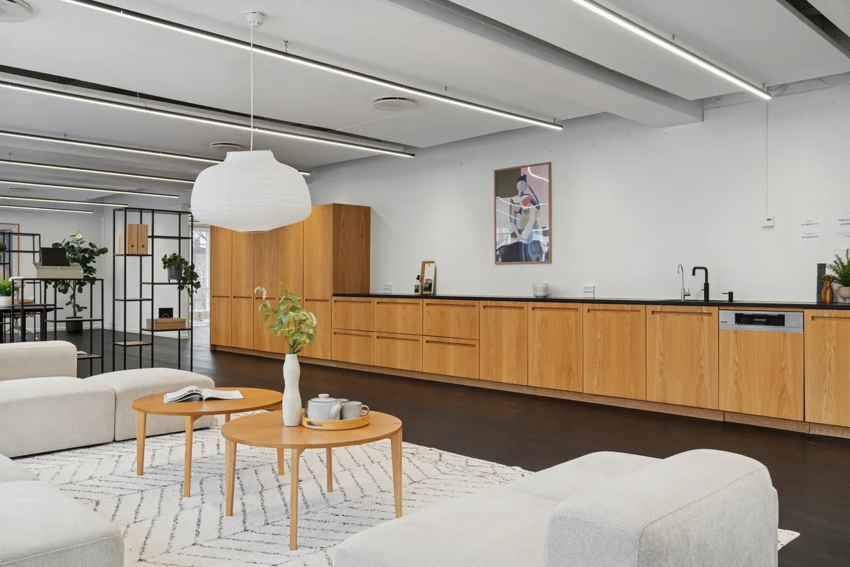 Kontor med åbent, moderne design og lyse farver. To bord med plante og tavle. Køkken med moderne møbler og lyse loftslampe. Sofas og hjørnebord i lys grå farve