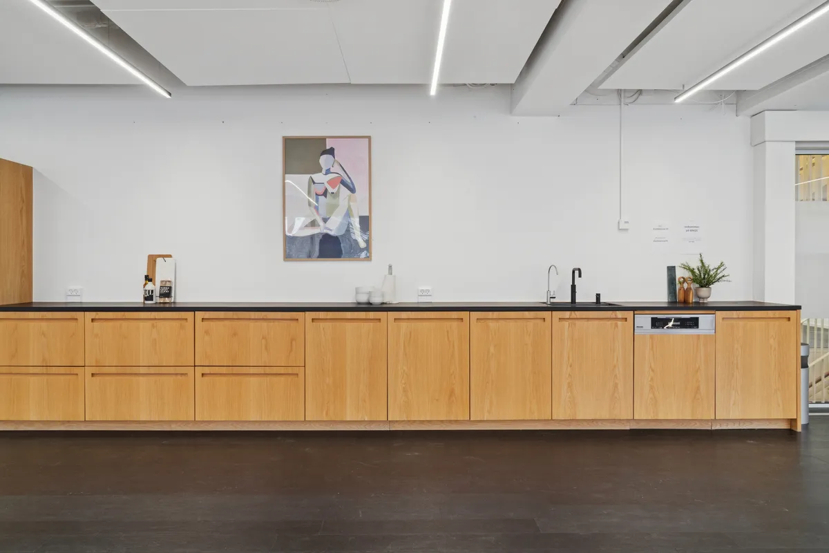 En moderne køkken i et kontor med lyse bøgetræsskabe og en sort granit bordplade med køkkenvask og opvaskemaskine. Væggen er hvid med en maleri i pastelfarver og lyse lysstriper.