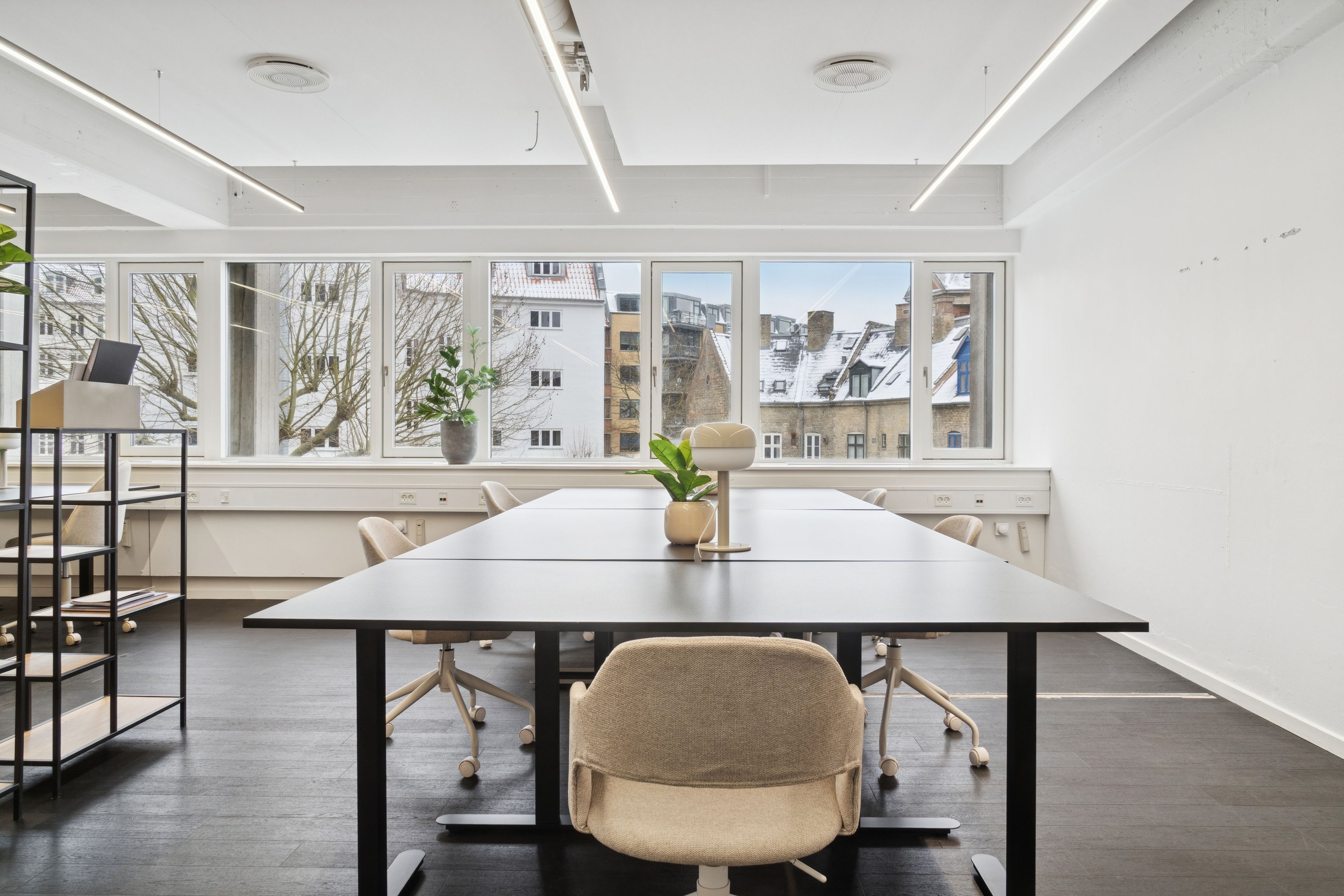 Moderne kontor med langt bord, høje ruder og lyse loftslampe, hvor sne dækker træer og bygninger udenfor