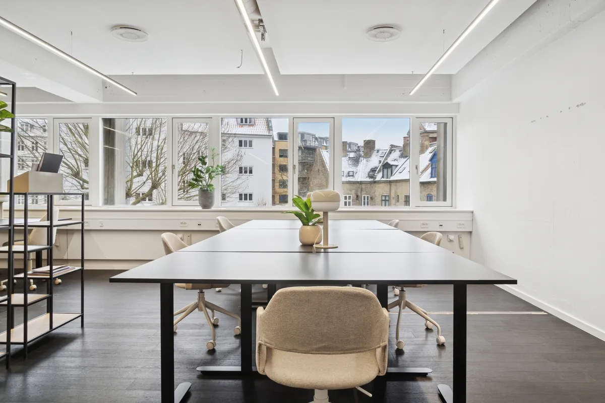 Moderne kontor med langt bord, høje ruder og lyse loftslampe, hvor sne dækker træer og bygninger udenfor