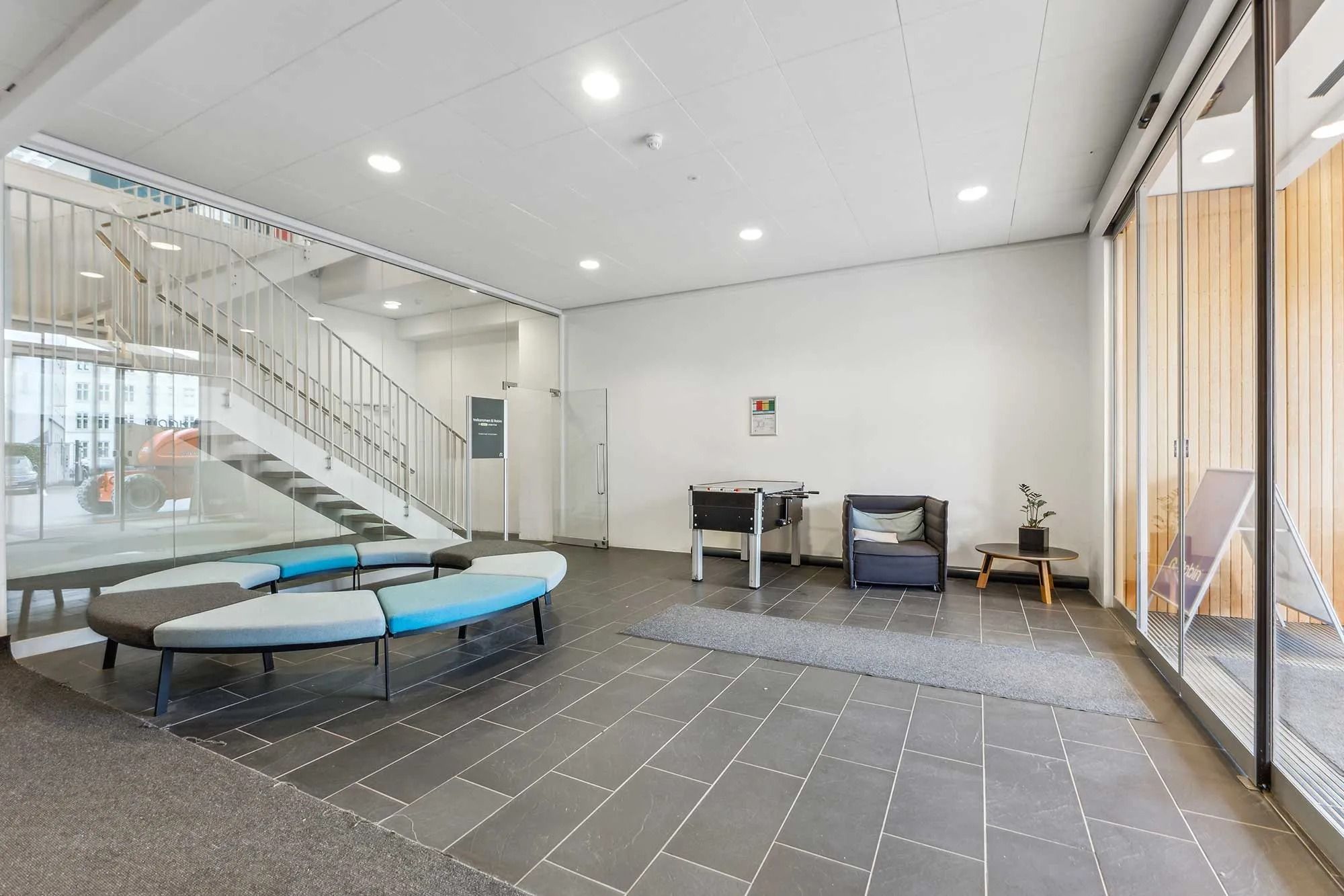 Indgangshallen med glasdøre, lysekroner, trappe, bord med plante, bord med fodboldbord og hvid sofa i moderne kontorbygning