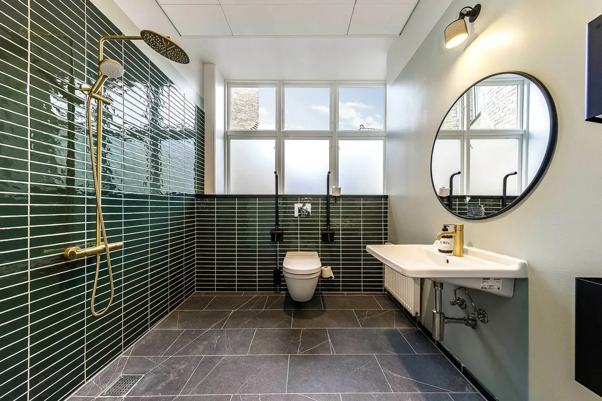 Toilette med toilet, rundt spejl, radiator, vindue, grønne fliser, toiletpapir og håndsæbe.