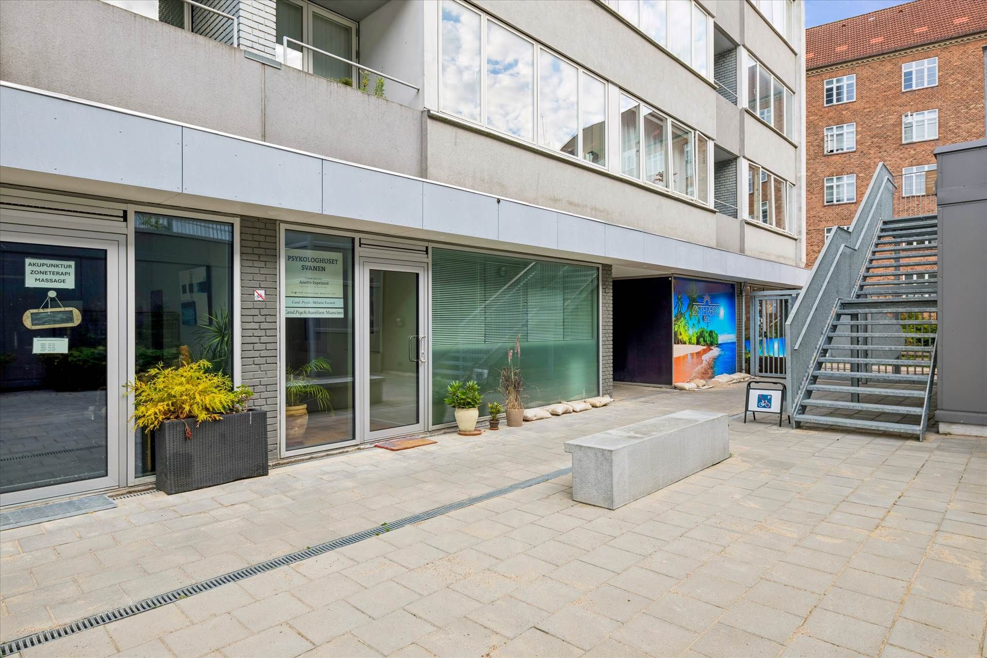 Udenfor klinik med to indgange, terrasse, grønne plantekasser og en trappe med skråning.