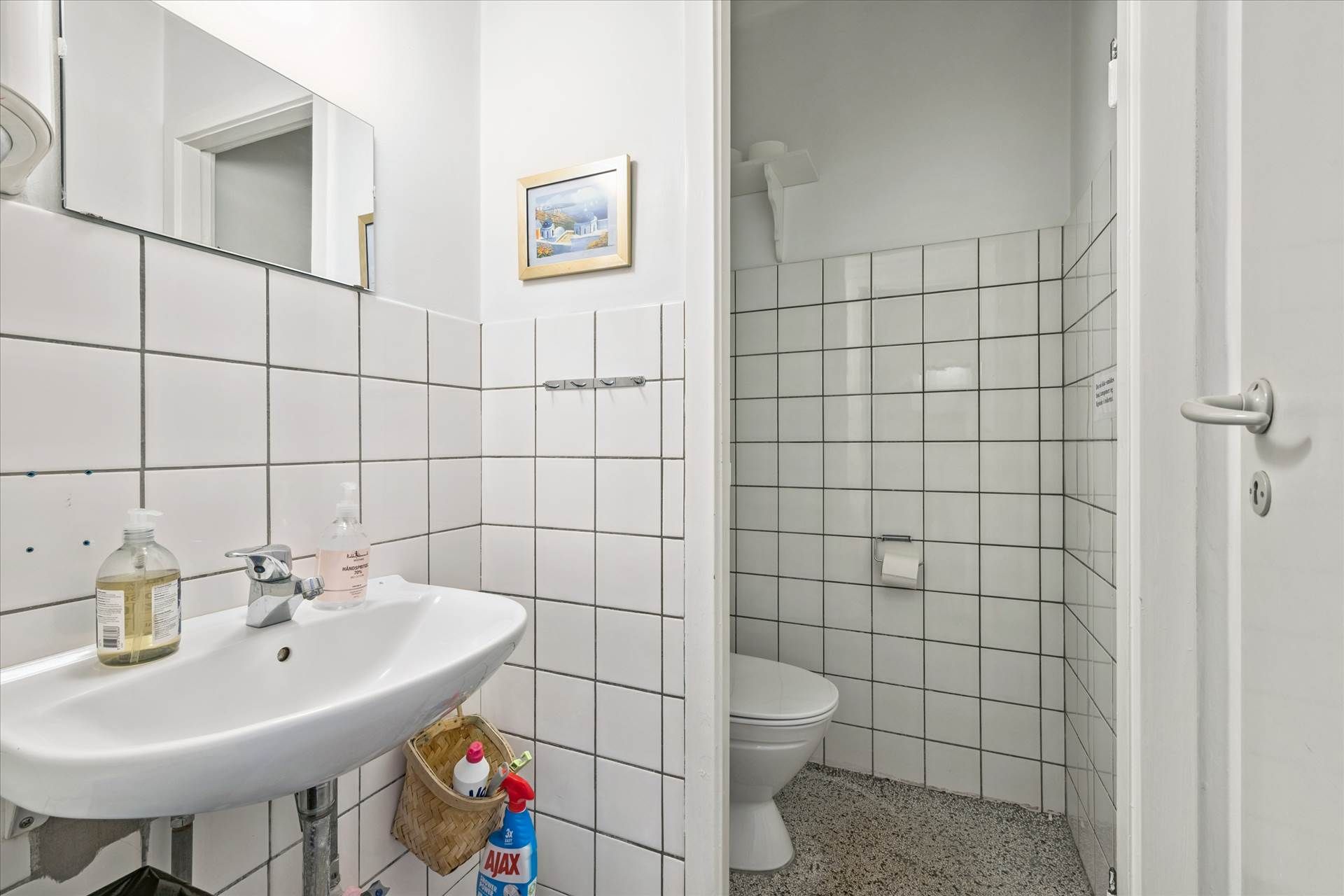 Hvidt badeværelse med toilet, håndvask, spejl og toiletpapir. Der er lysegrå flise på væggene.