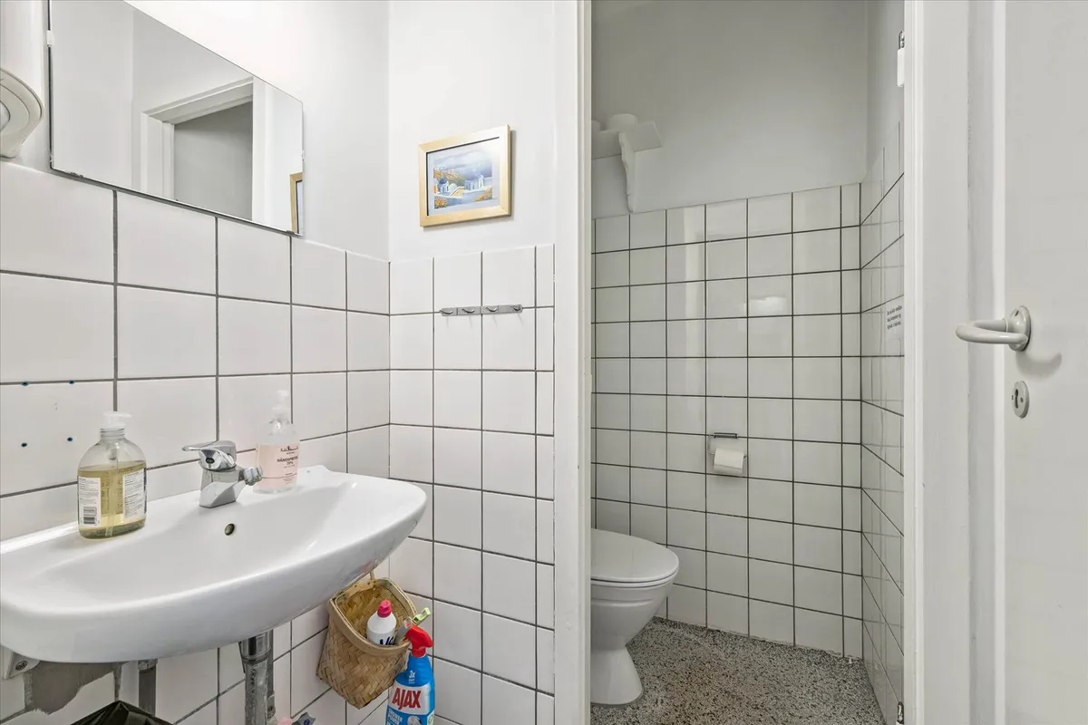 Hvidt badeværelse med toilet, håndvask, spejl og toiletpapir. Der er lysegrå flise på væggene.