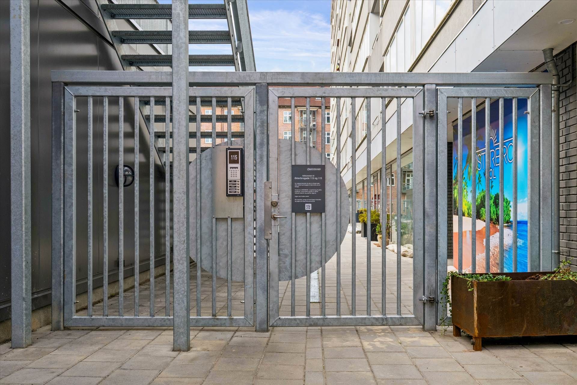 En stålporte med åbningsmekanisme og plakat udenfor en bygning med skrånende tag og facade med tekst og billede