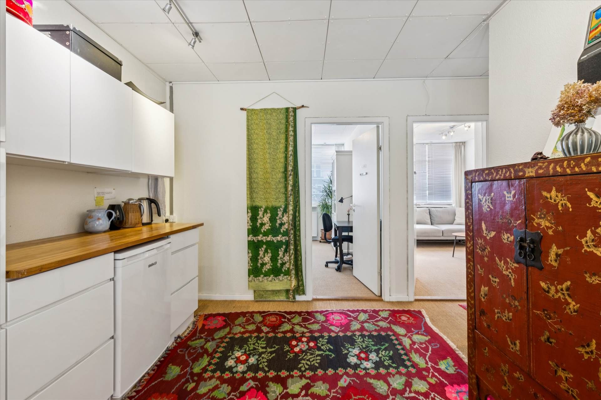 Køkken med hvide skabe og træbordplade, tebrygger, kaffemaskine og en stor orientalsk tæppe på gulvet