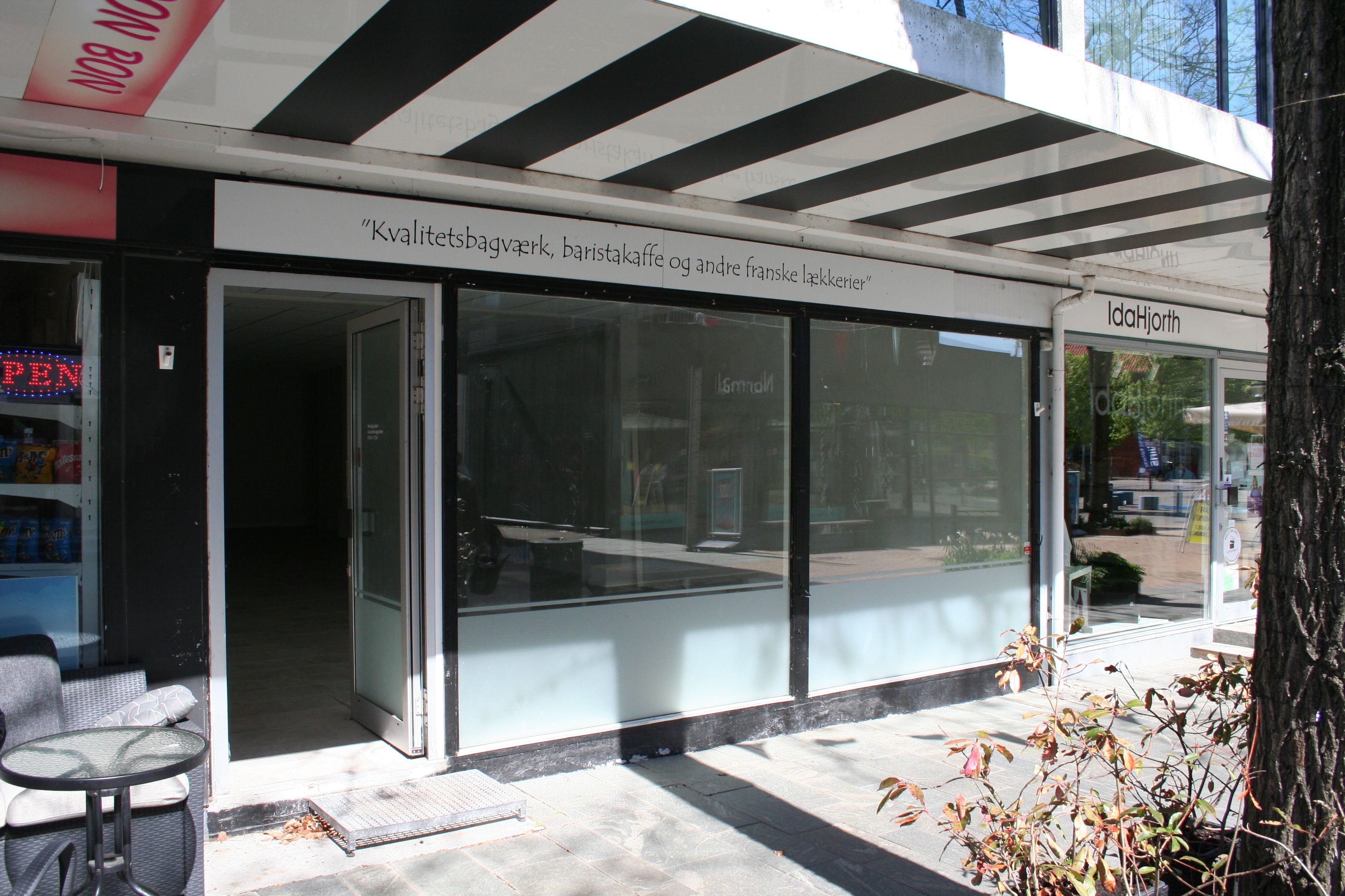 Forretningslokaler med indgang og vinduer samt en lille spisestue med bord og stole. Butikken har en skrift med