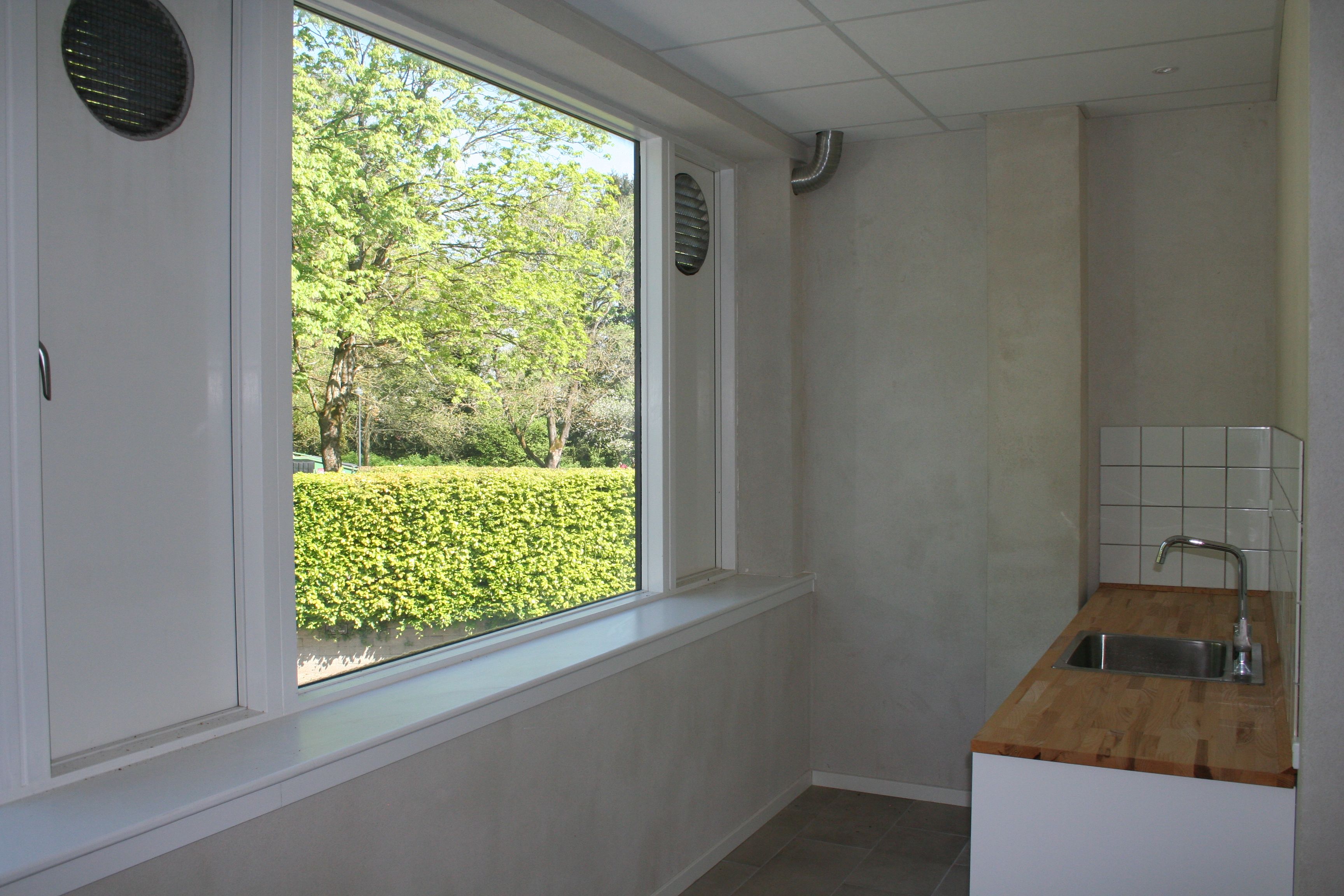 En rummelig stue med lyse gulve og hvide vægge, et vindue med udsigt til en grøn buskhed og hvide kakelover på væggen ved