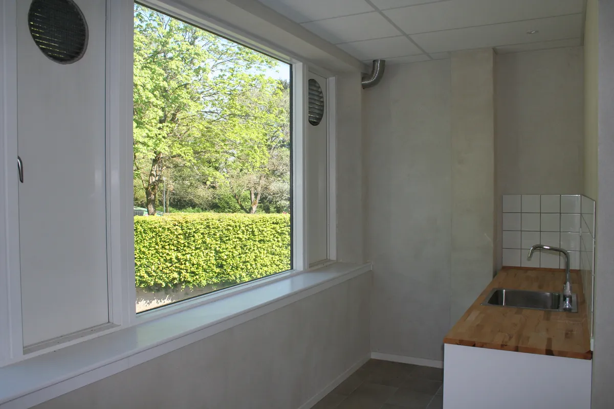 En rummelig stue med lyse gulve og hvide vægge, et vindue med udsigt til en grøn buskhed og hvide kakelover på væggen ved