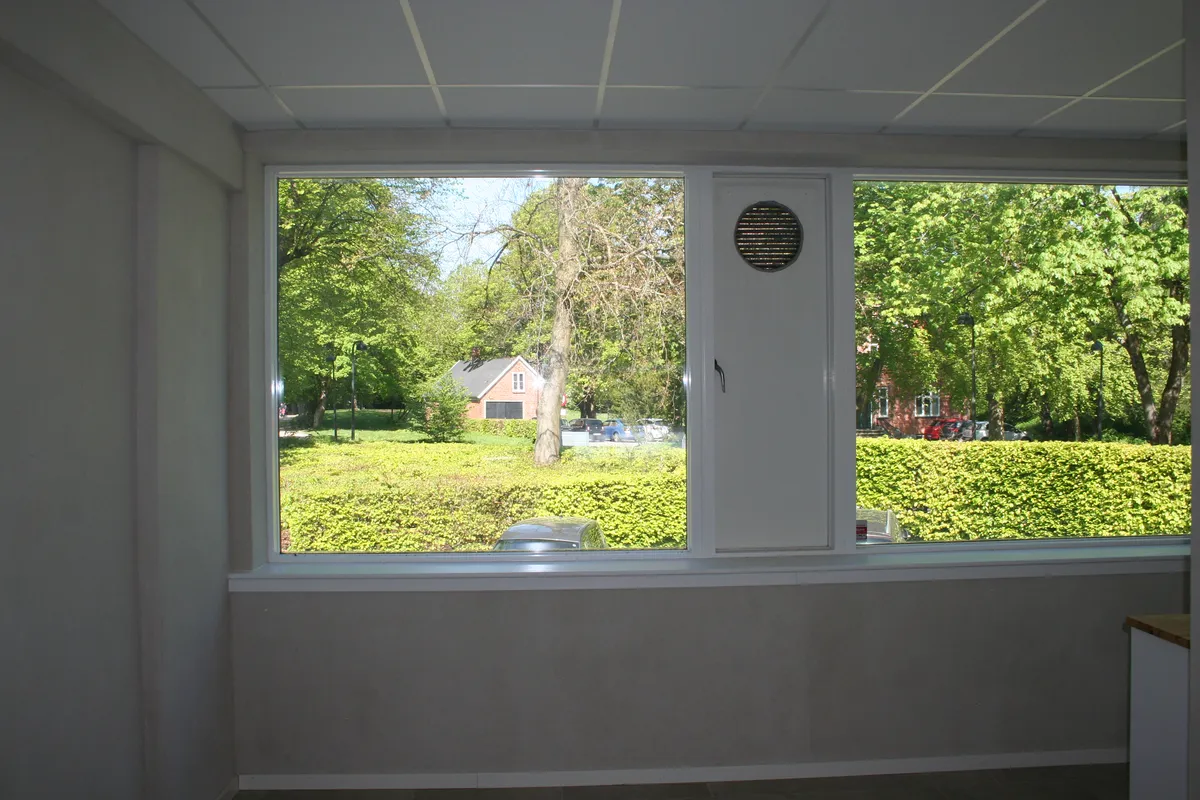 Vindue i hvidt rum med udsigt til gård med grønne buske og parkeringsplads med biler og hus med rød tagpanne.