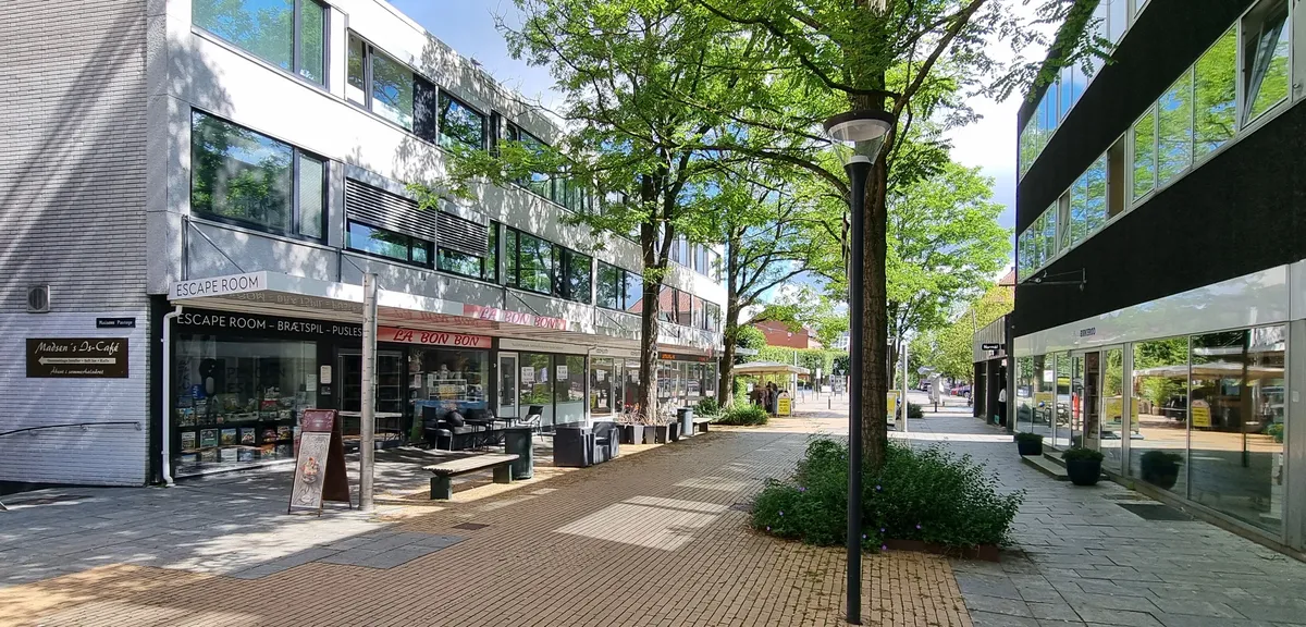 Gågade med høje bygninger og grønne træer, med søjler, bænke og forretninger med skilte som Escape Room og La Bon Bon