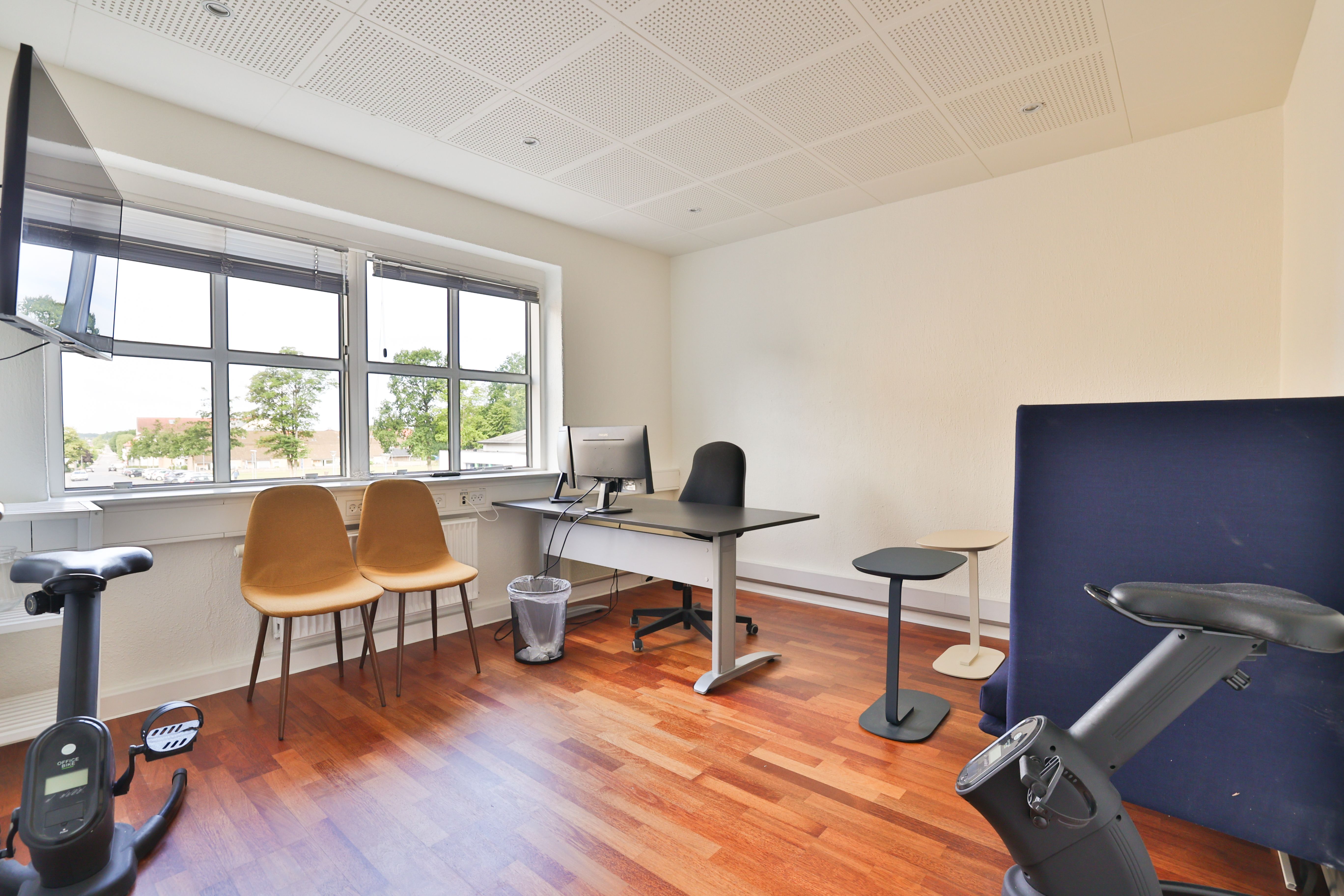 Kontor med tre lysestole, to skriveborde, skrivebordsstol, tv og motionscykel i en gulvbelægning med lyse toner