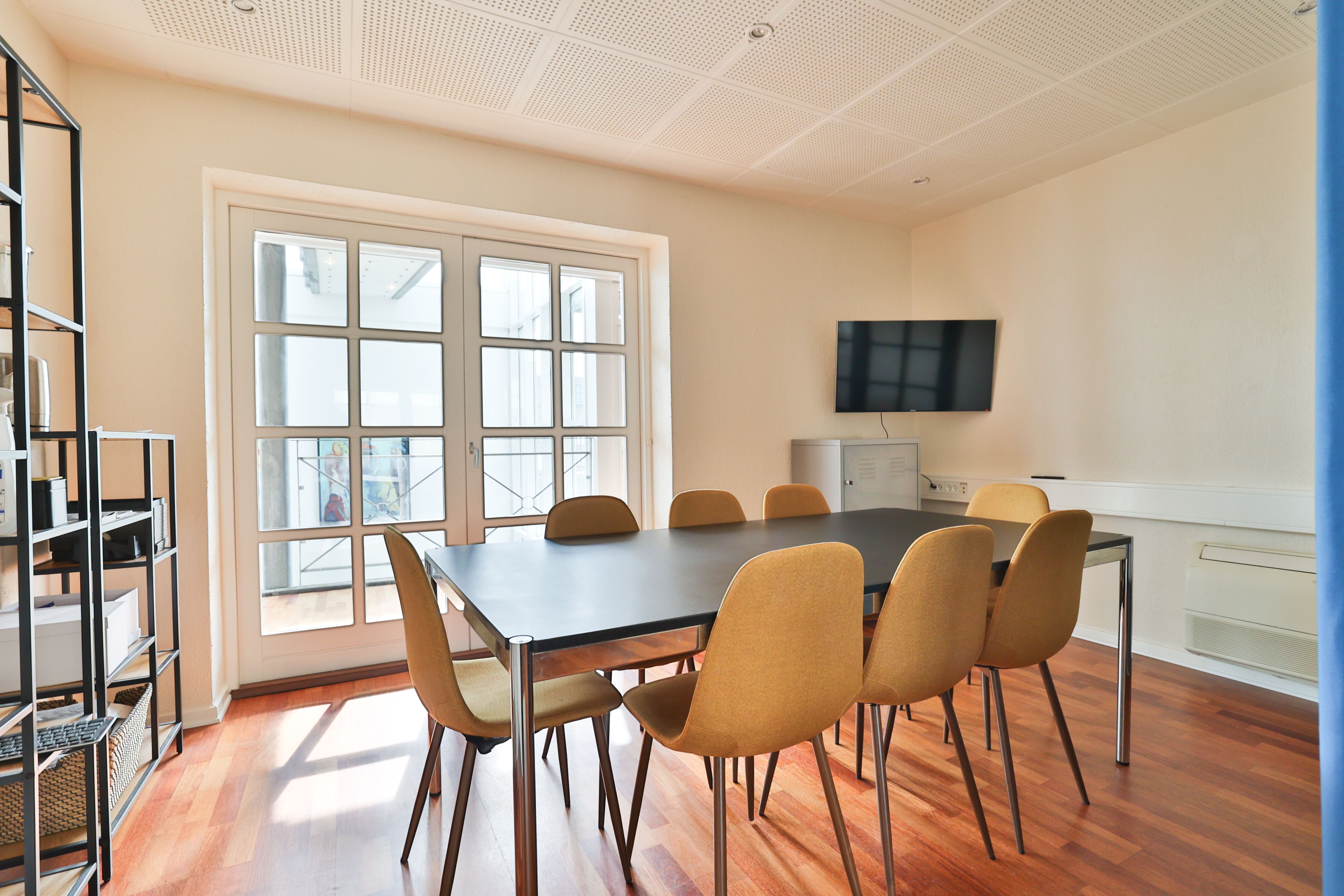 Et kontorlokale med en stor bord, flere stole og en tv-skærm på væggen. Loftet er lavet af hvide paneler og gulvet er lavet