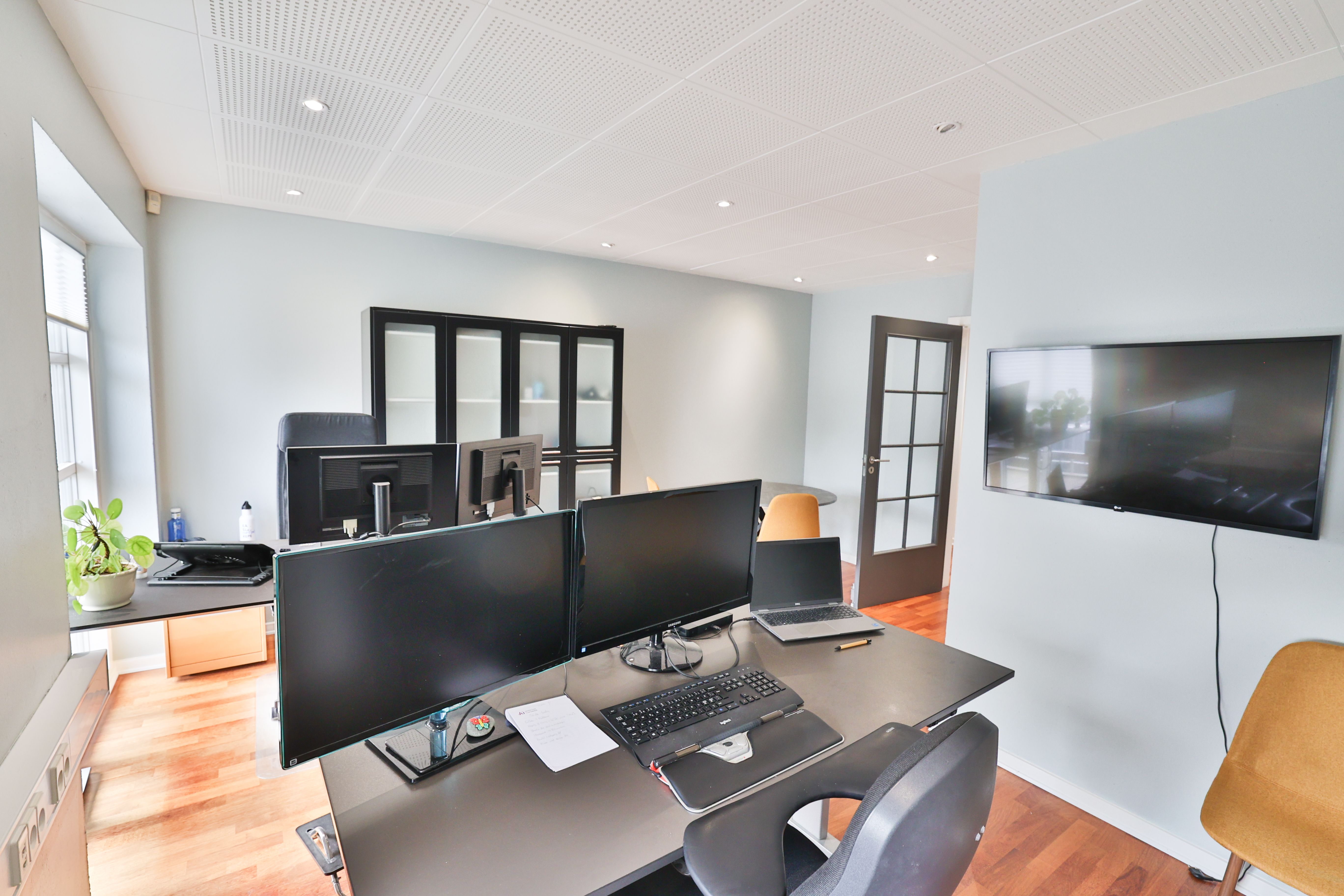 Kontor med to stående skriveborde, hver med to skærme, skrivebordslys og computermus samt en lukket dør med vinduesruder