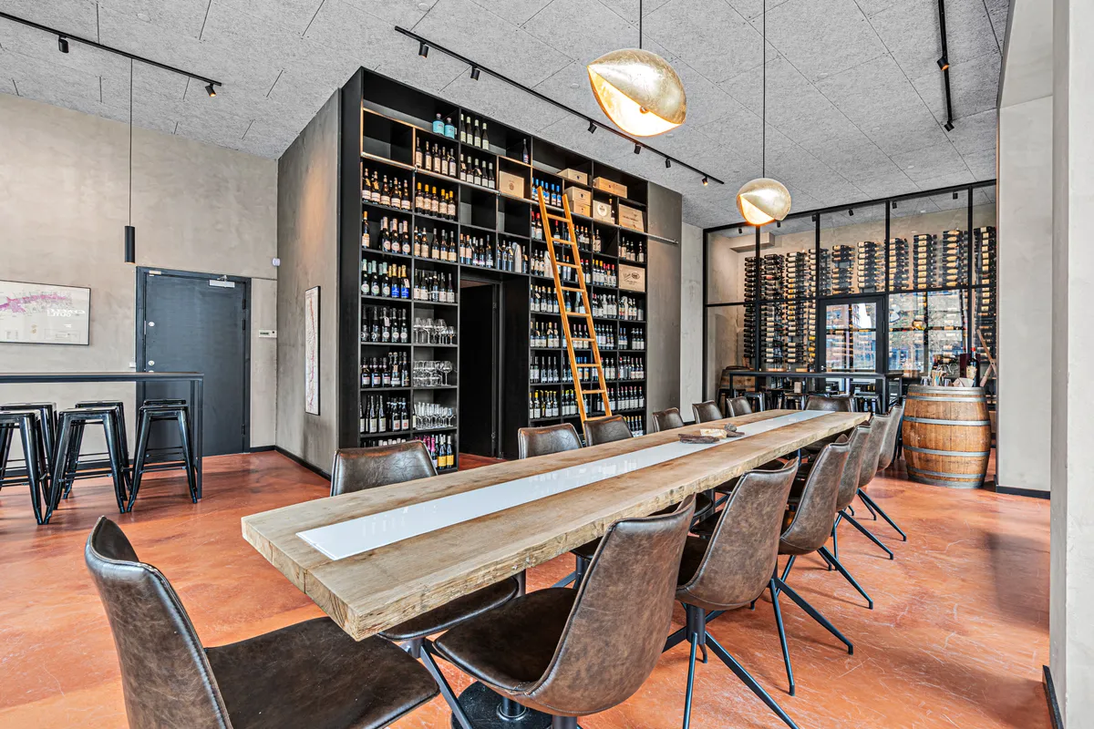 Lang træbord i moderne vinbar med læderstole og store vinreoler. Gul stige og to lysekroner i loftet.