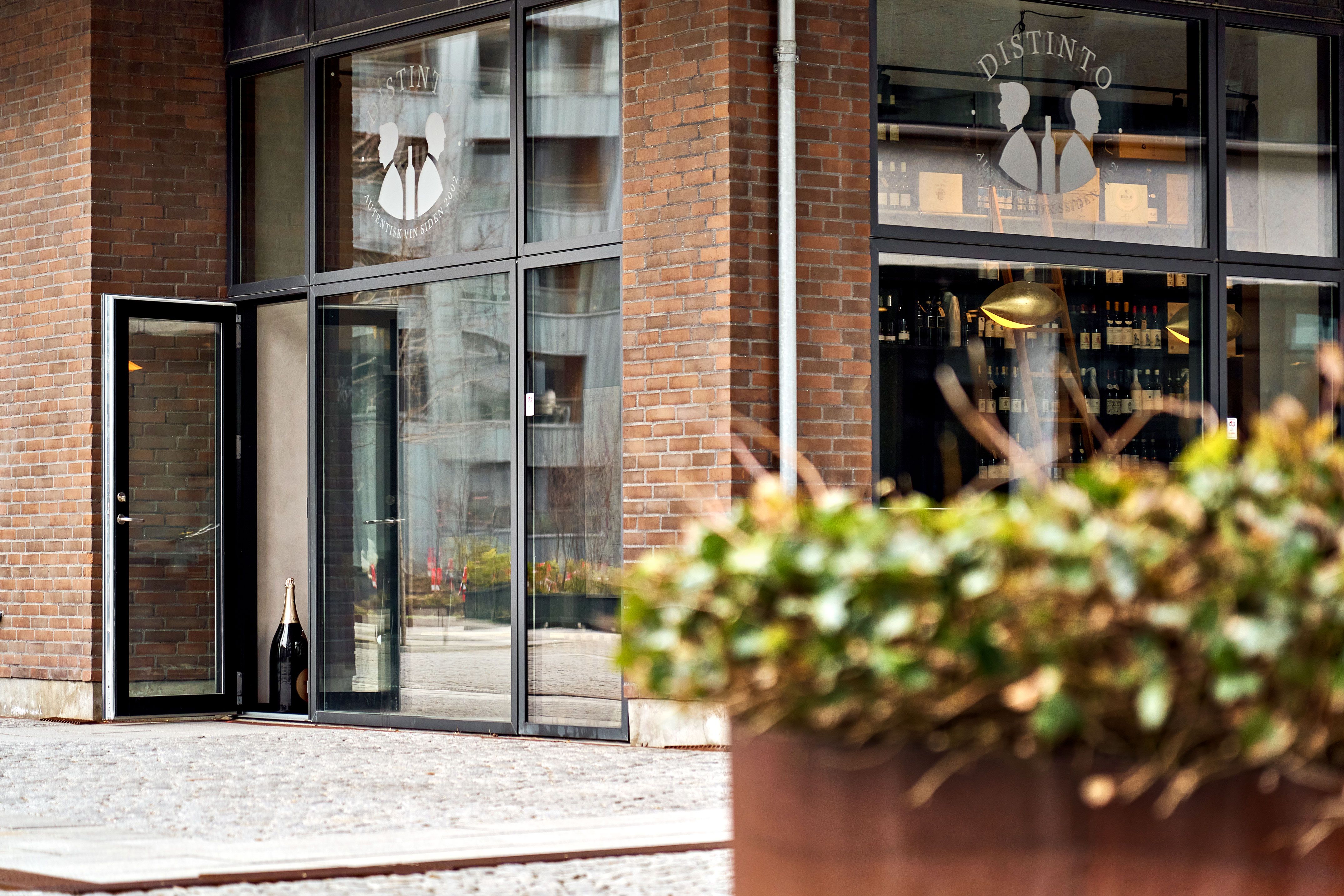 Foto af Facaden af en vinbar med glasdøre og et vinflaske logo i det ene vindue og en stor plantepotte foran indgangen